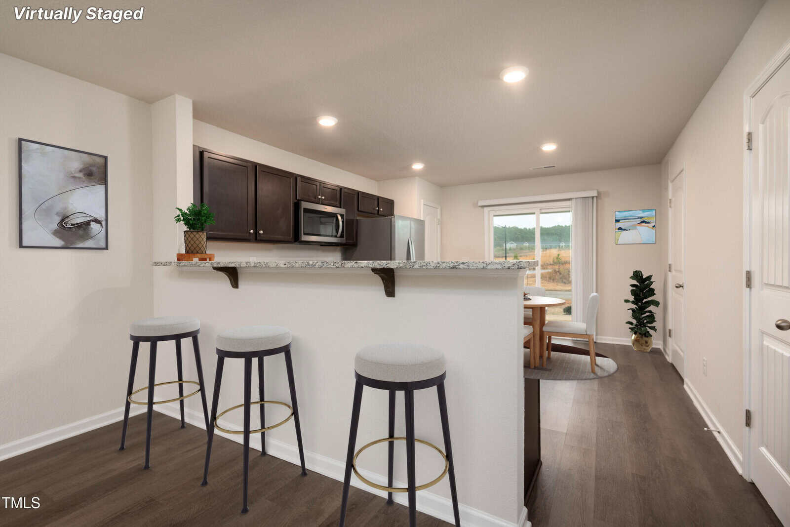 A modern kitchen with dark cabinets, a granite breakfast bar with three stools, stainless steel appliances, and wood floors; a dining area with a round table is visible in the background near sliding glass doors.