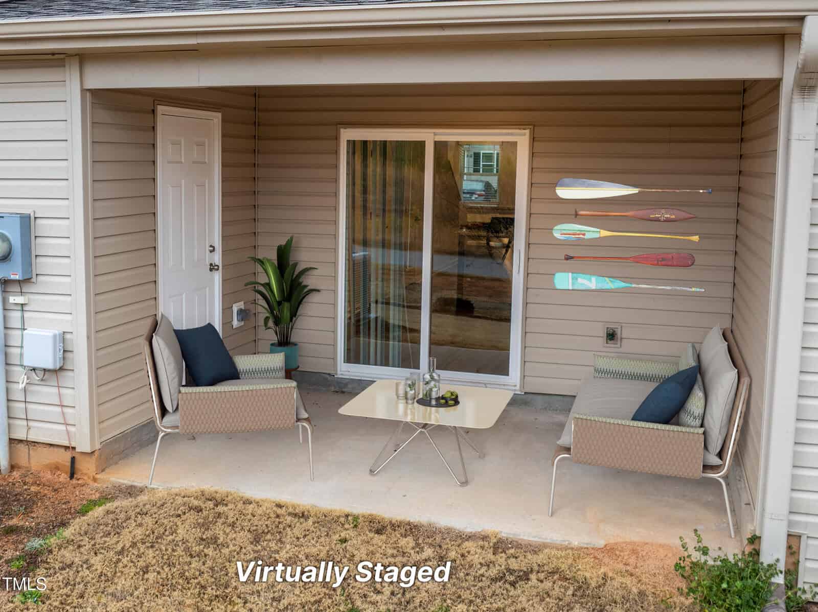 A small covered patio with tan siding features two cushioned chairs, a glass-top table, a potted plant, and four decorative paddles mounted on the wall. The image is labeled "Virtually Staged.