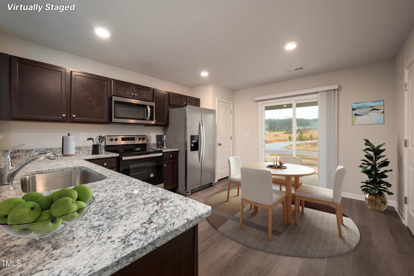 A modern kitchen with dark wood cabinets, stainless steel appliances, and granite countertops. A small round dining table with four chairs is set on a rug near sliding glass doors, overlooking an outdoor view. "Virtually Staged" is written in the corner.