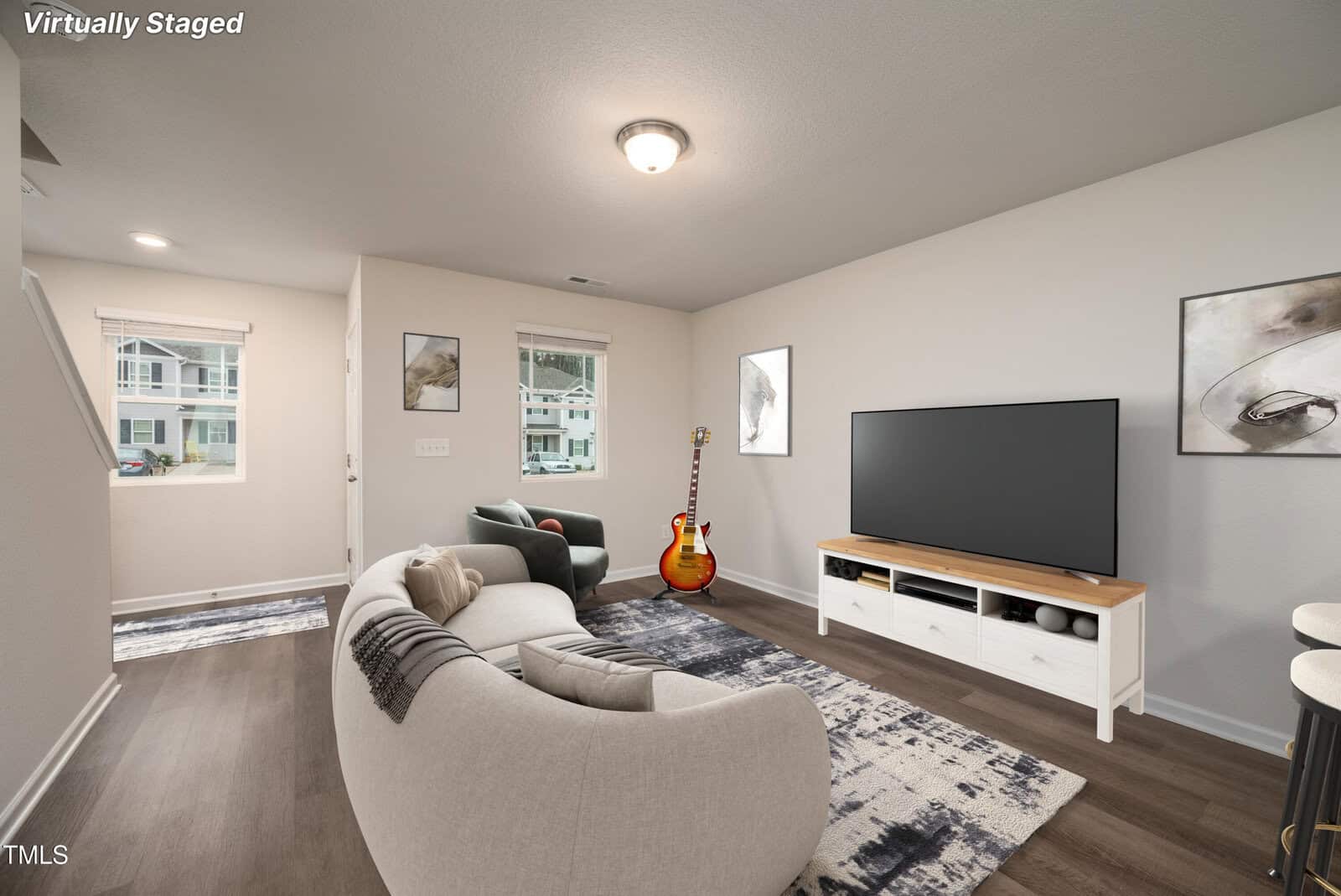 A living room with a curved sofa, a TV on a white stand, a guitar, and two abstract wall art pieces. Two windows let in natural light. The floor is wood, and a rug is under the sofa. The image is virtually staged.