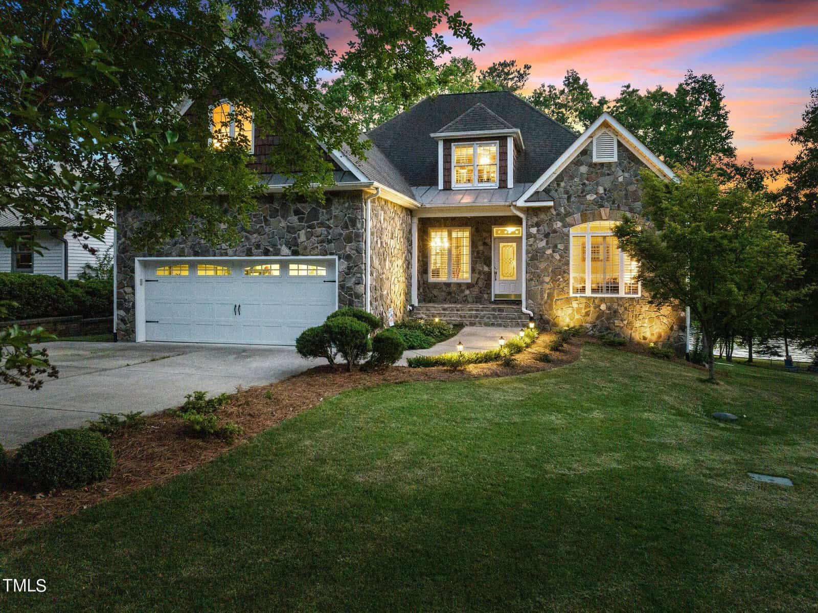 Stone house with a two-car garage, large windows, and illuminated entryway at dusk. Lush green lawn, landscaped bushes, and mature trees surround the home under a colorful sunset sky.