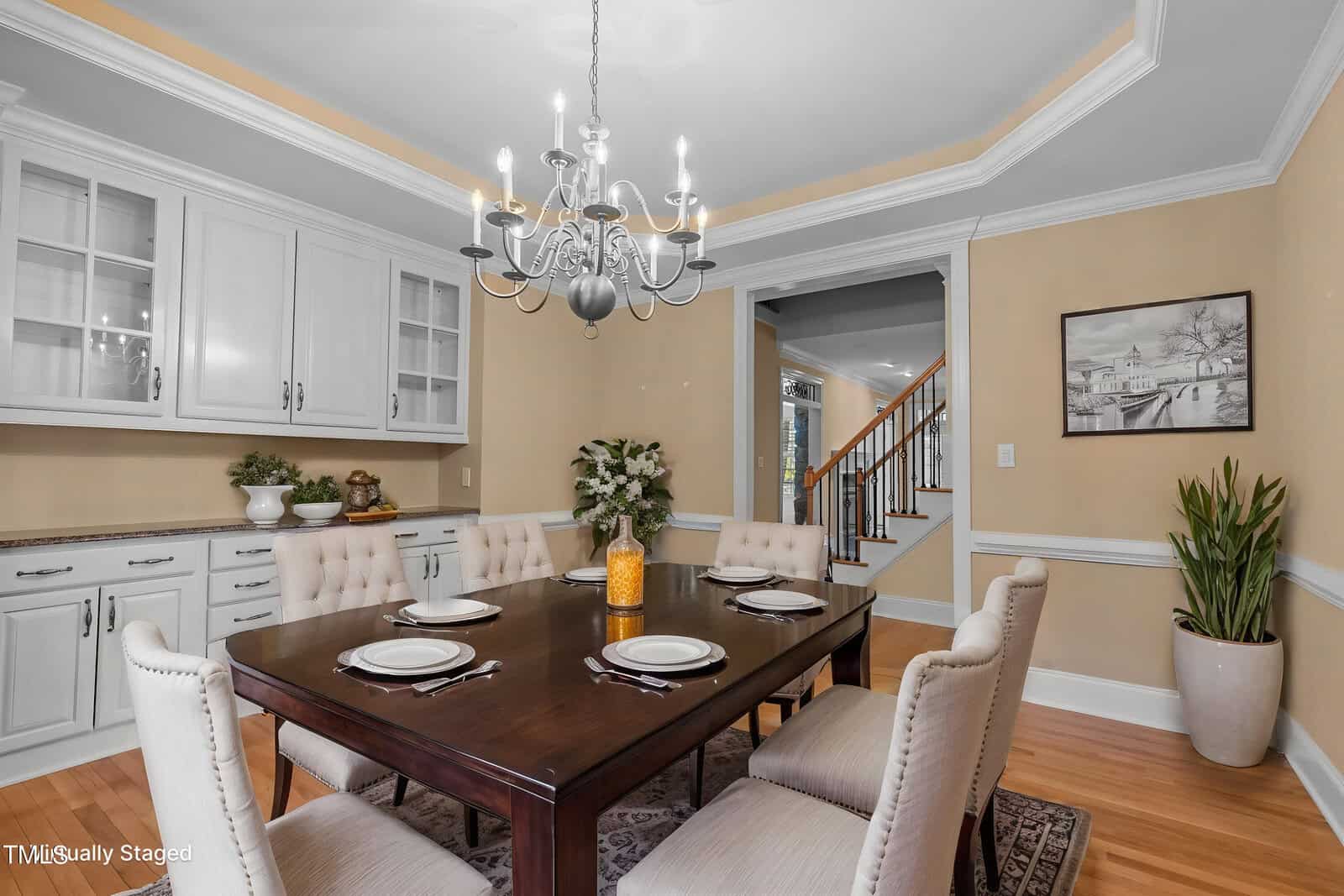A dining room with a dark wood table set for six, beige upholstered chairs, a chandelier, white cabinetry, potted plants, and framed art on tan walls. A staircase is visible through an open doorway in the background.