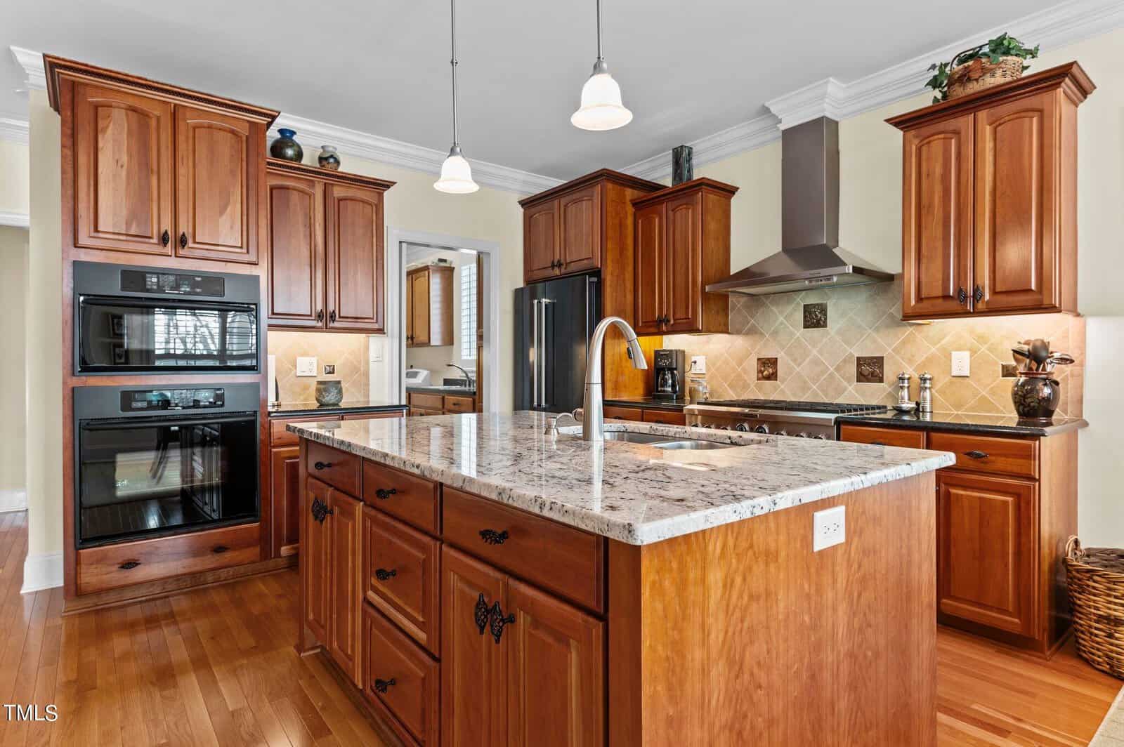 Spacious kitchen with wood cabinets, granite island with sink, stainless steel appliances, double oven, range hood, tile backsplash, and pendant lights.