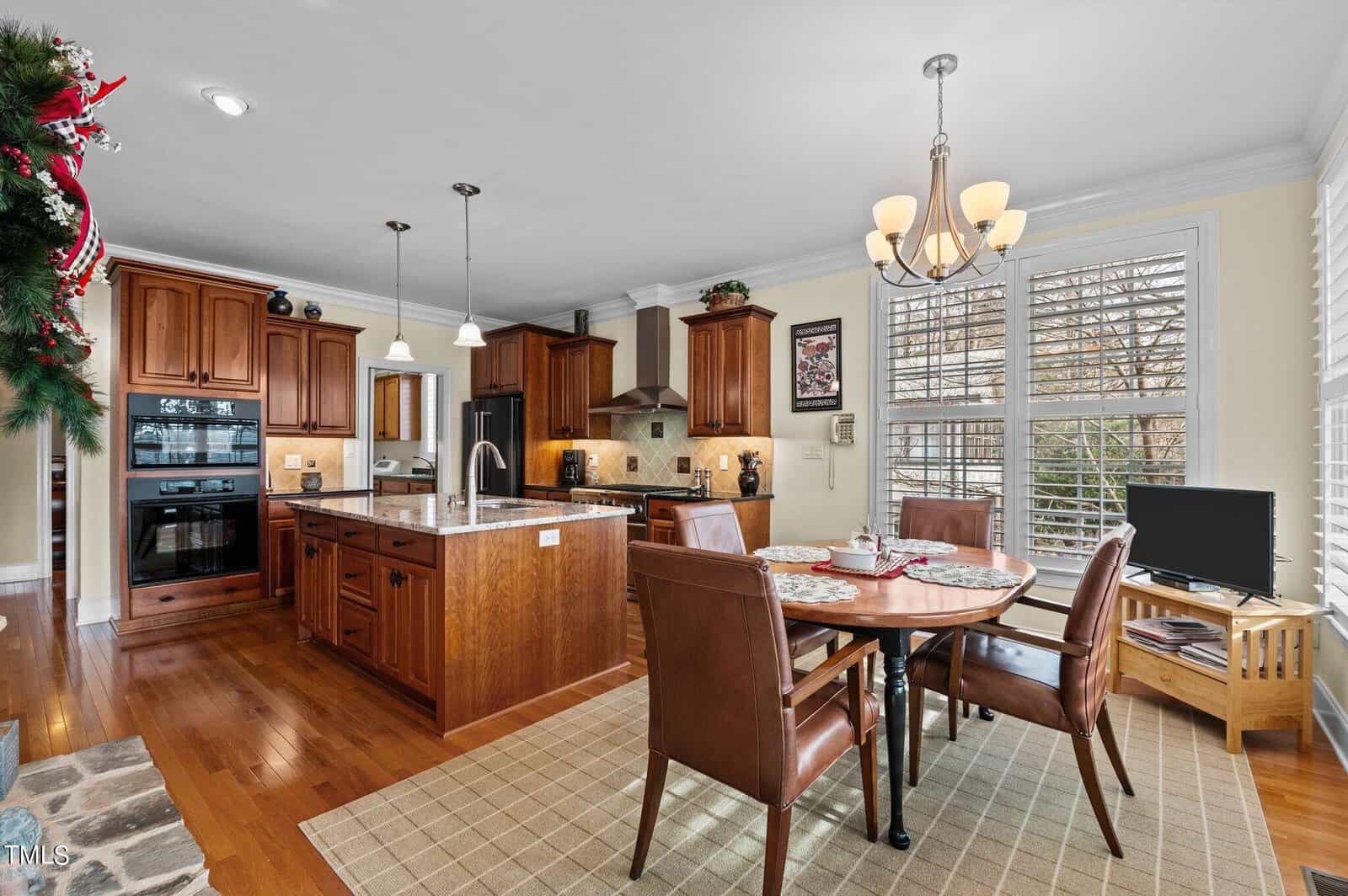 Spacious kitchen and dining area with wooden cabinets, island with sink, dining table set for four, pendant lights, chandelier, and large windows with plantation shutters, all featuring warm, cozy decor.