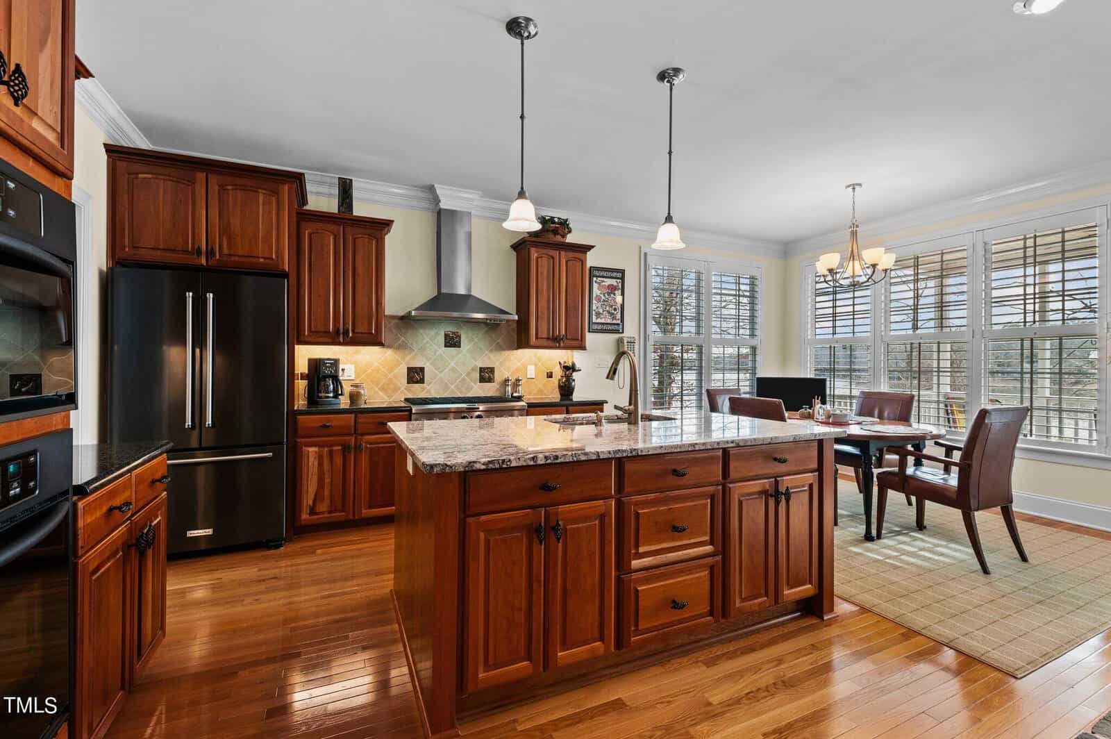 Spacious kitchen with dark wood cabinets, granite countertops, stainless steel appliances, and an island. A dining area with a table and four chairs is next to large windows with white shutters.