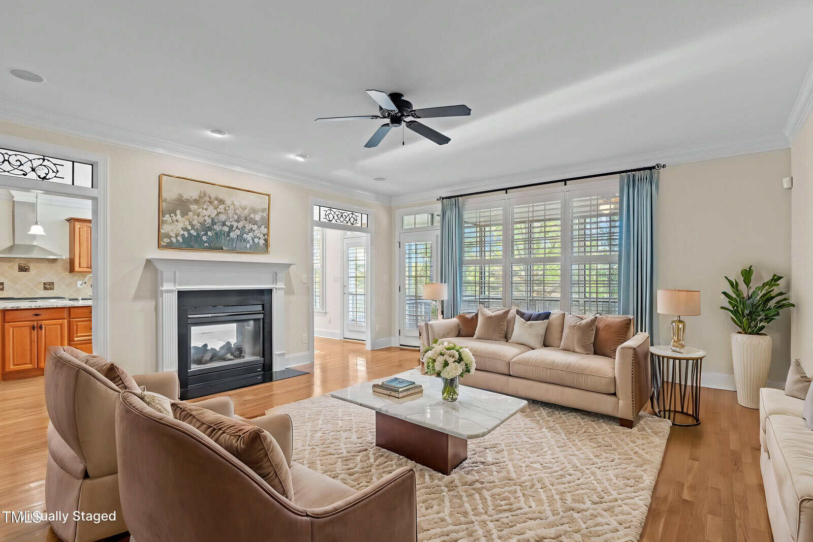 Bright living room with two beige sofas, a marble coffee table, an area rug, modern armchairs, a fireplace with artwork above, large windows with blue curtains, hardwood floors, and a ceiling fan. Kitchen visible in background.