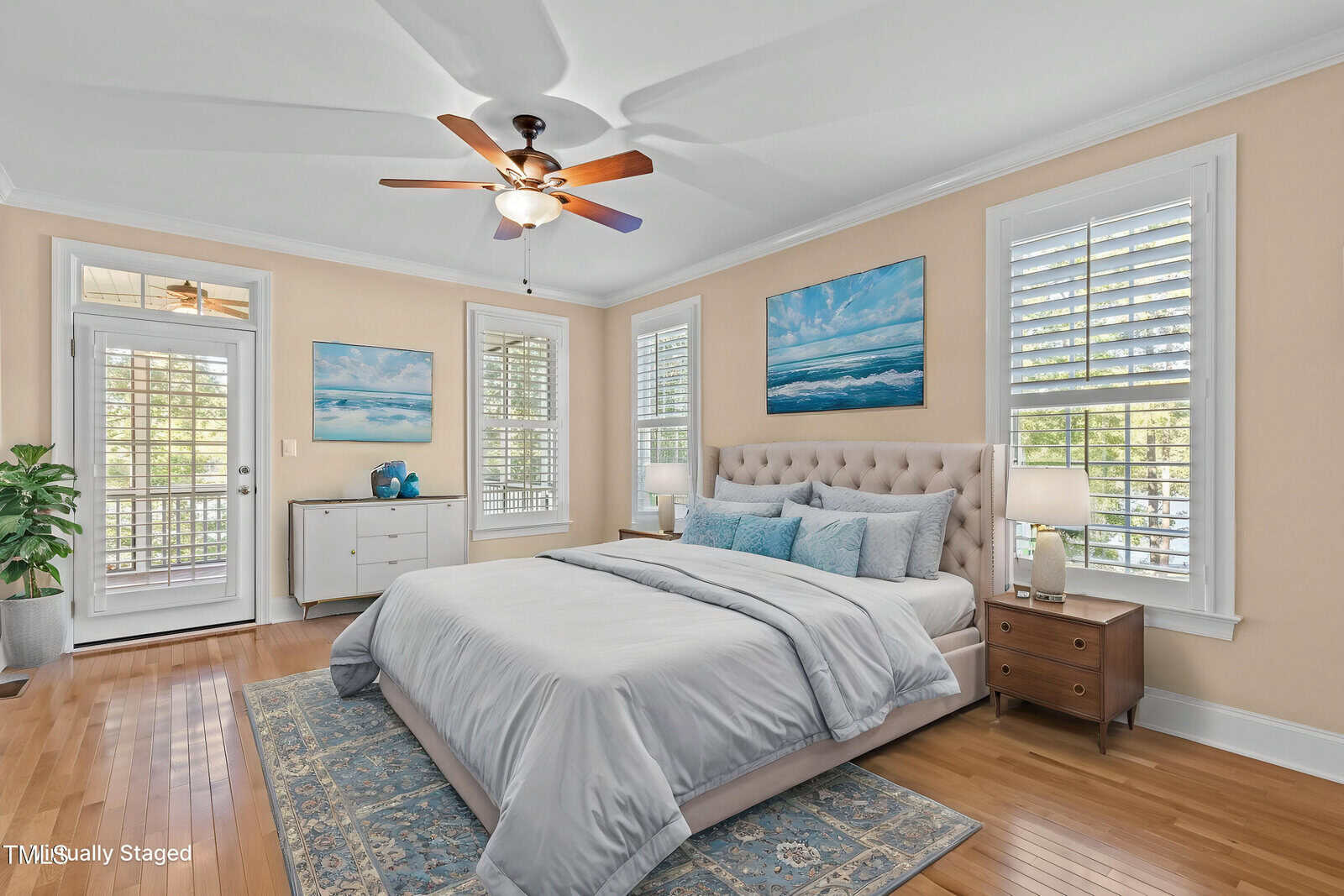 A bright, airy bedroom with tan walls, wood floors, and white trim. The room features a large bed with light bedding, two nightstands, a dresser, ocean-themed art, and several windows with white shutters.