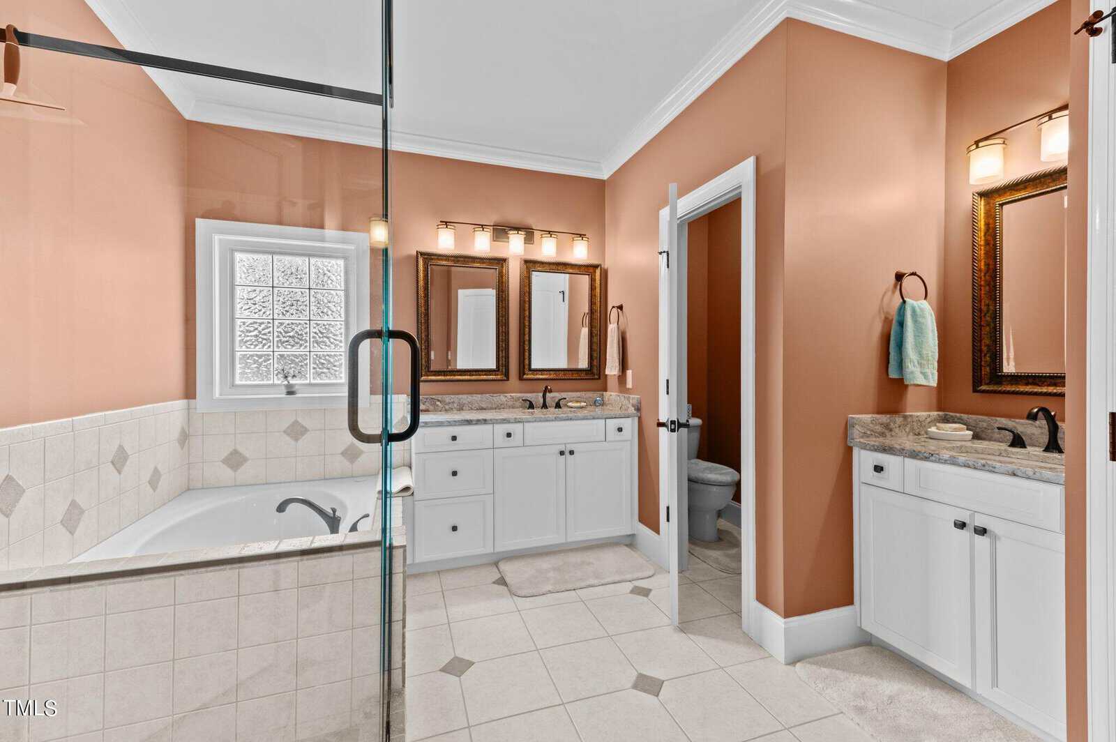 Spacious bathroom with peach-colored walls, white tiles, a built-in bathtub, double vanity with marble countertops and mirrors, glass shower, and a separate toilet area with a towel hanging on the wall.