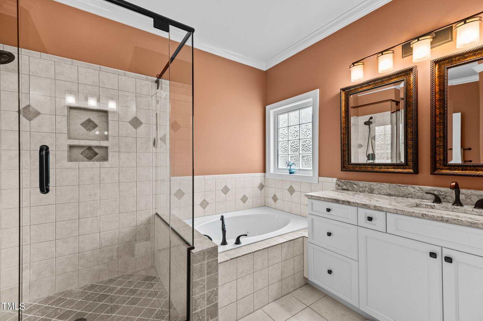 A bathroom with light brown walls features a glass-enclosed shower, a built-in bathtub with beige tiles, a white double-sink vanity with granite countertop, and two framed mirrors under three wall lights.