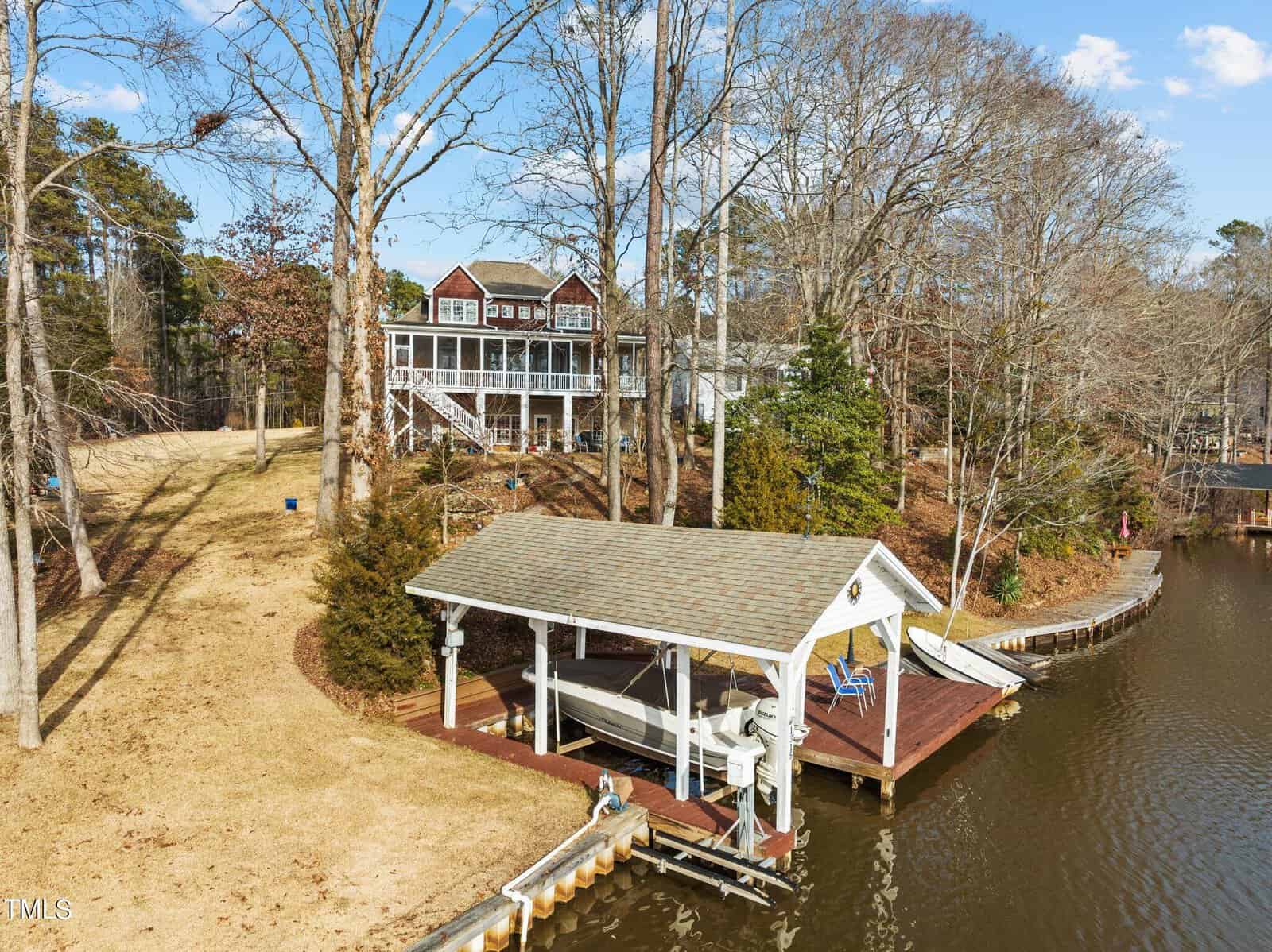A large house with multiple decks overlooks a quiet lake. In the foreground, a covered dock with a boat and chairs sits at the water's edge, surrounded by bare trees and a sloping lawn on a clear day.