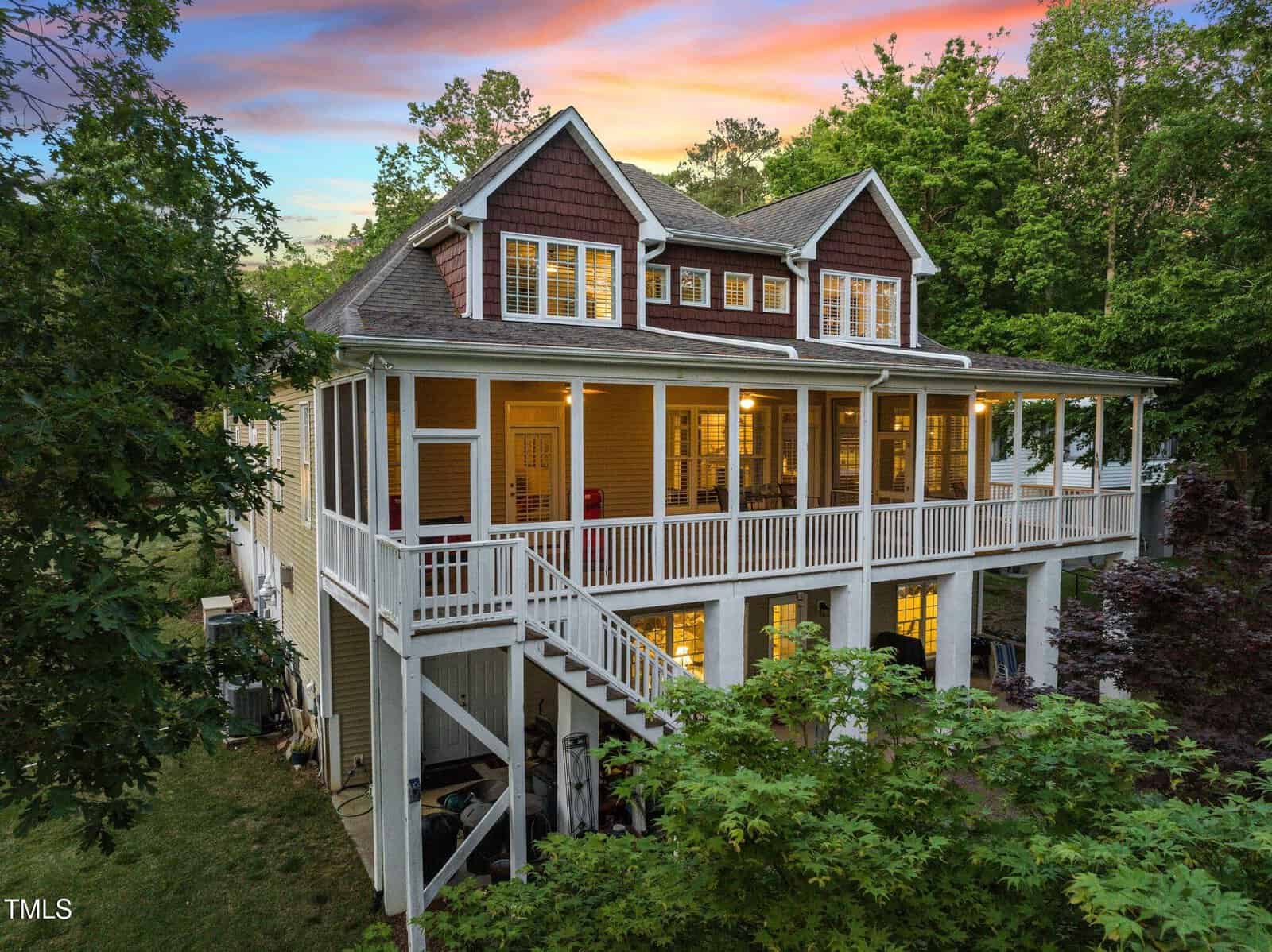 A two-story house with a large screened-in porch, lit warmly at dusk. The home features brown and white siding, a gabled roof, and an exterior staircase, surrounded by lush green trees.