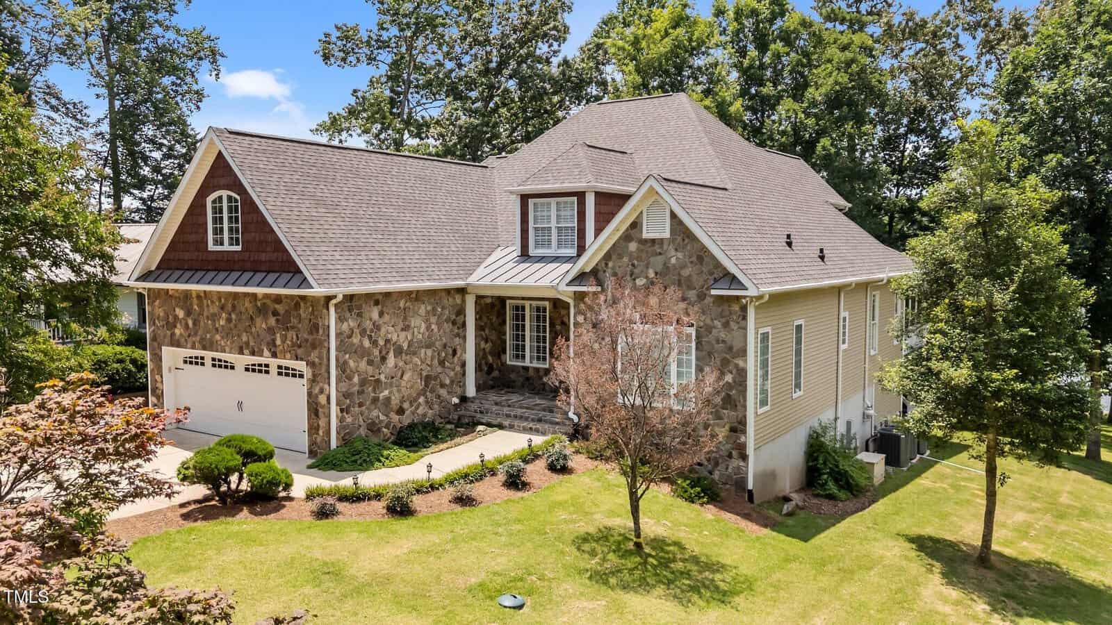 A large two-story house with stone and beige siding, a gabled roof, and an attached two-car garage, surrounded by a well-maintained lawn, trees, and landscaping on a sunny day.