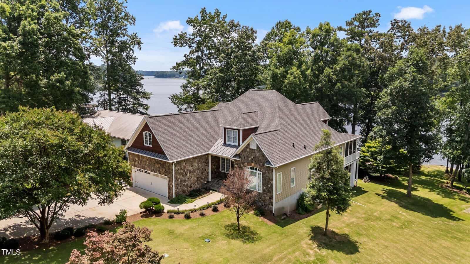 Nestled at 1315 Sagamore Drive, Louisburg, this large two-story home with a stone and brick exterior sits on a grassy lawn surrounded by trees, overlooking a lake under a blue sky with scattered clouds.