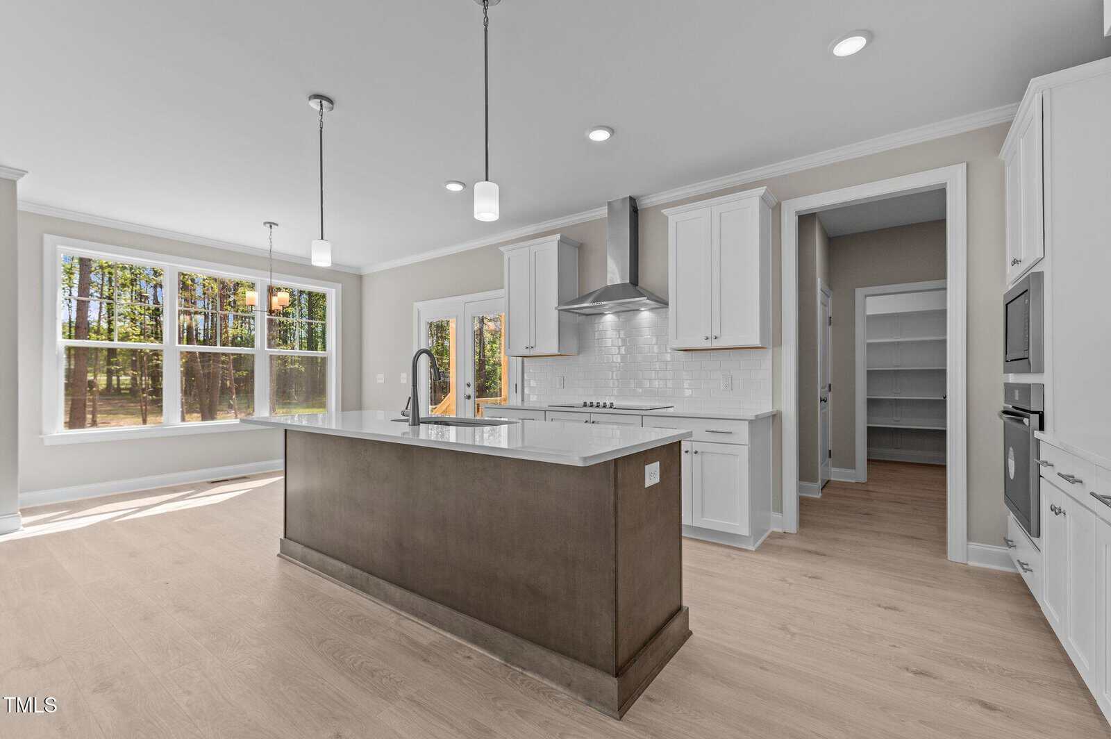 Modern kitchen with white cabinets, stainless steel appliances, a large island with dark wood base, light wood floors, pendant lights, and a walk-in pantry. Large windows provide natural light and a view of trees outside.