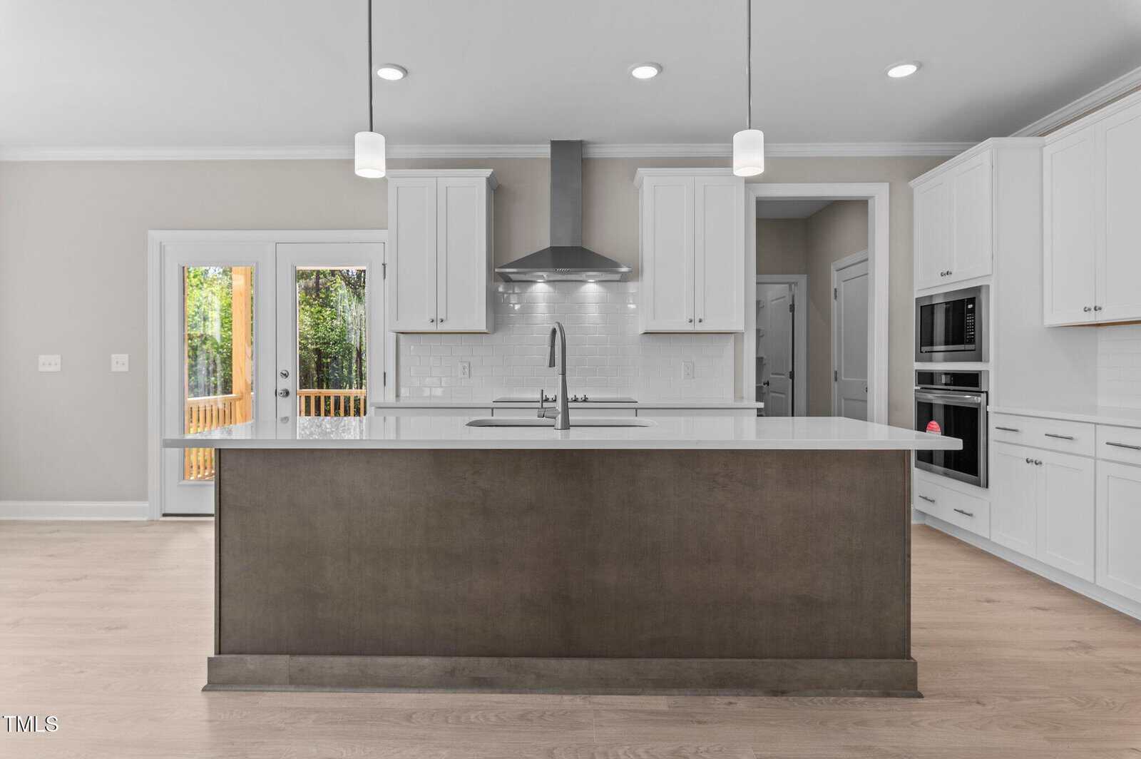 Modern kitchen with a large island, white cabinets, stainless steel appliances, pendant lights, a double oven, and glass doors leading to a patio. The space features light wood floors and neutral walls.
