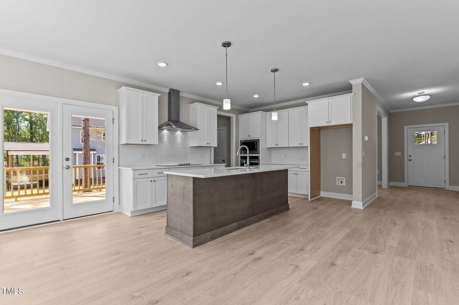 Modern kitchen with white cabinets, stainless steel appliances, a center island with pendant lights, light wood floors, and double doors leading to a deck. Open layout with view to entryway and lots of natural light.