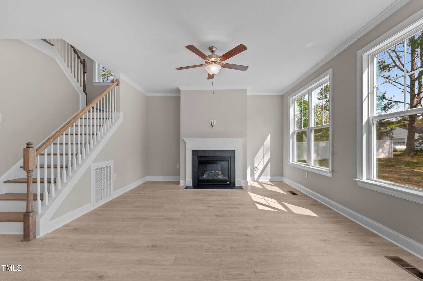A bright, empty living room with light wood floors, large windows, a white fireplace, ceiling fan, and a staircase with a wooden handrail on the left side. Sunlight streams through the windows.