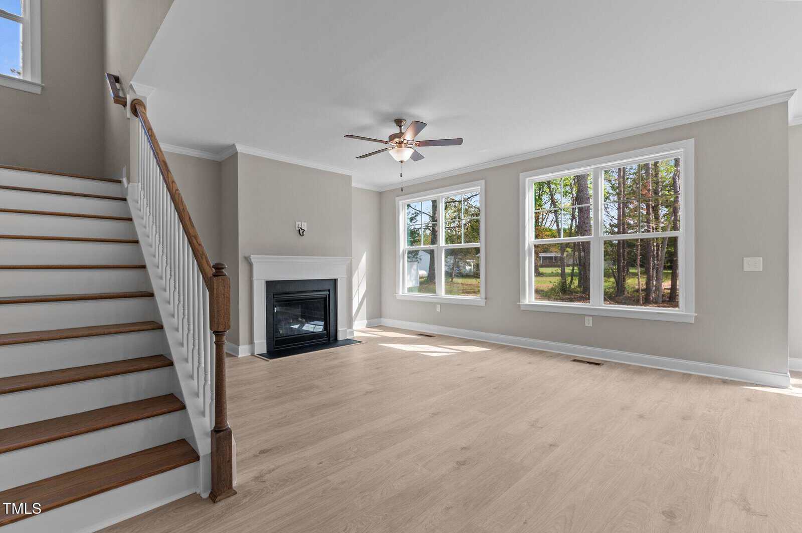 A bright living room with light wood floors, a white fireplace, large windows, a ceiling fan, and a wooden staircase leading upstairs.