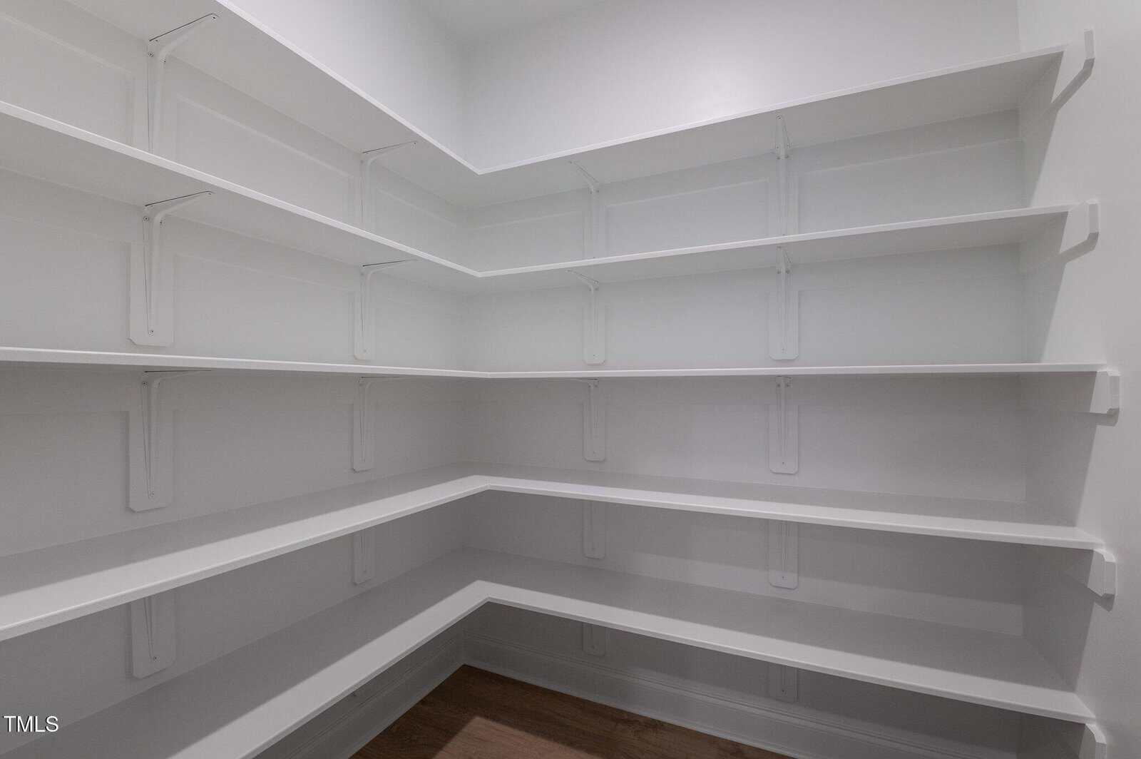 An empty walk-in pantry with white walls, multiple rows of white shelves lining the corners, and a wooden floor.