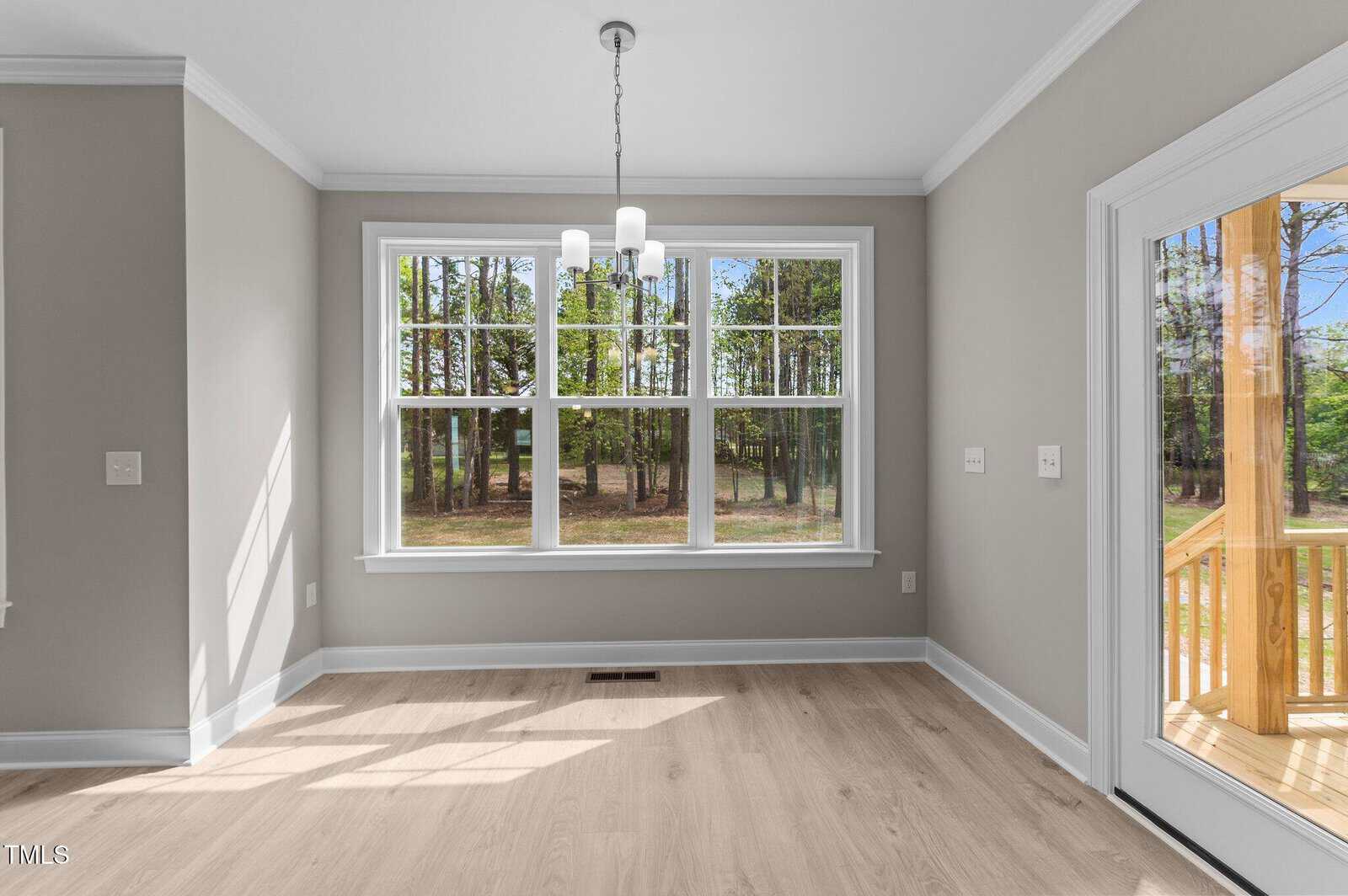 A bright, empty room with light wood floors, gray walls, a modern ceiling light fixture, and large windows offering a view of trees and a forested backyard. A glass door leads to a wooden porch.