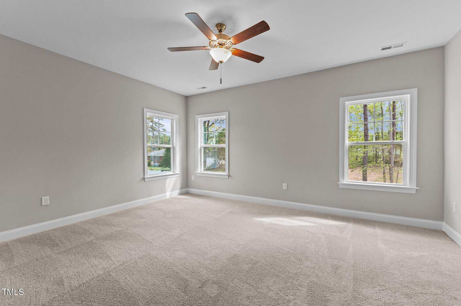 Spacious empty room with beige carpet, light gray walls, three windows, a ceiling fan with lights, and natural sunlight streaming in.