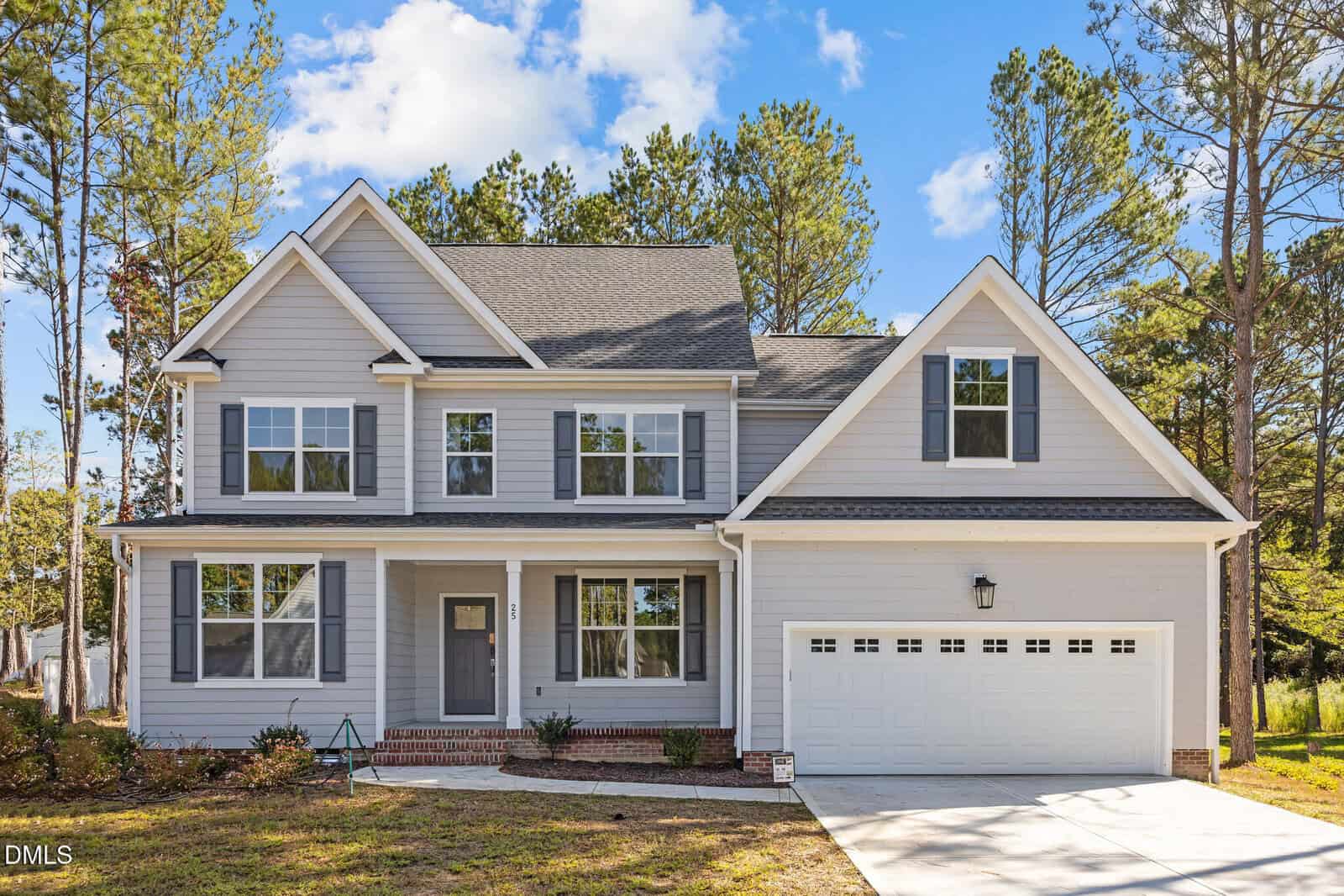 A two-story modern suburban house with gray siding, white trim, and shutters. The home has a covered front porch, multiple large windows, and an attached two-car garage, surrounded by tall pine trees and blue sky.