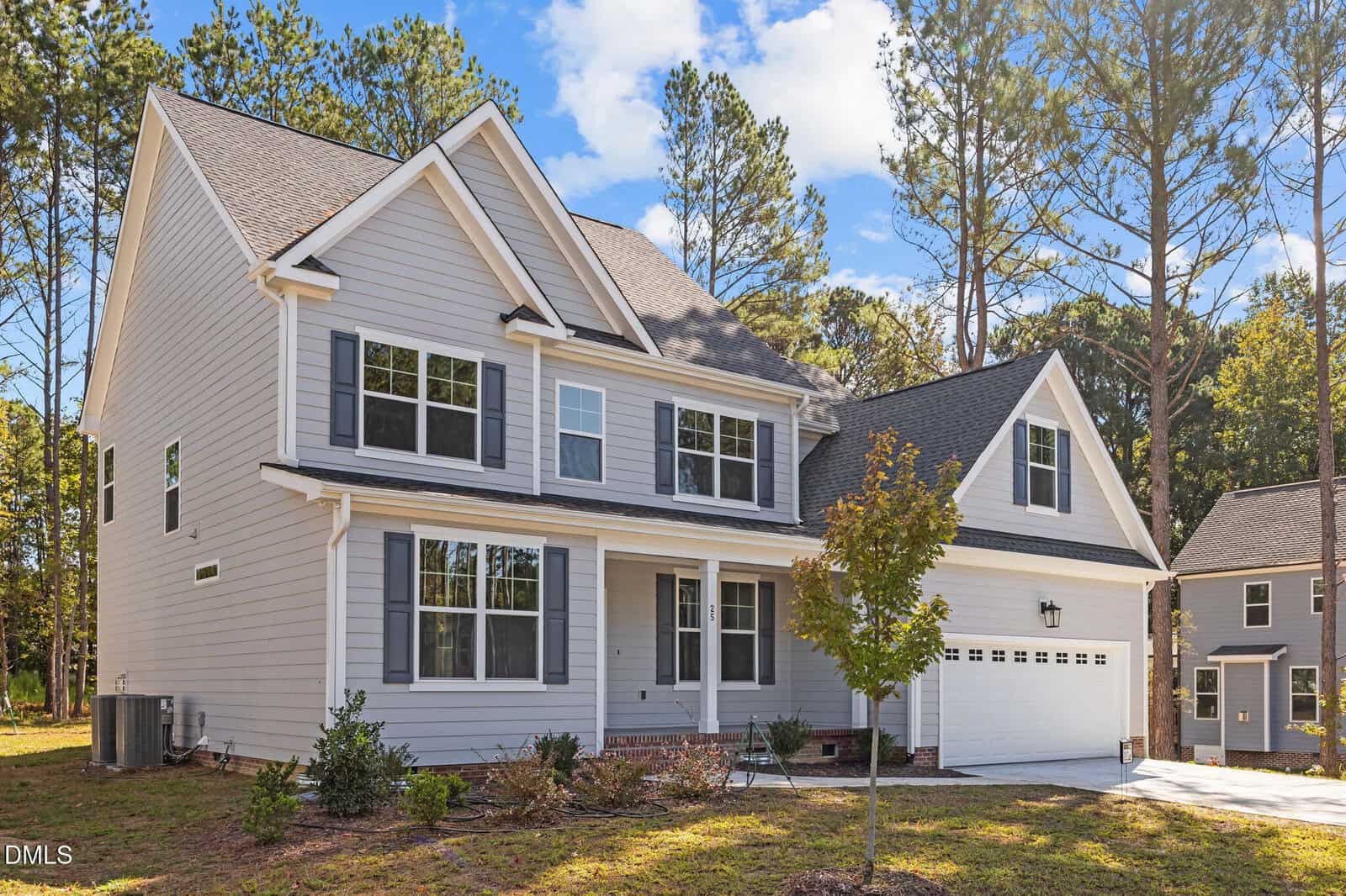 A two-story modern suburban house with gray siding, white trim, dark shutters, and a double garage. The front yard has a small tree, shrubs, and a brick walkway. Tall pine trees are in the background under a blue sky.