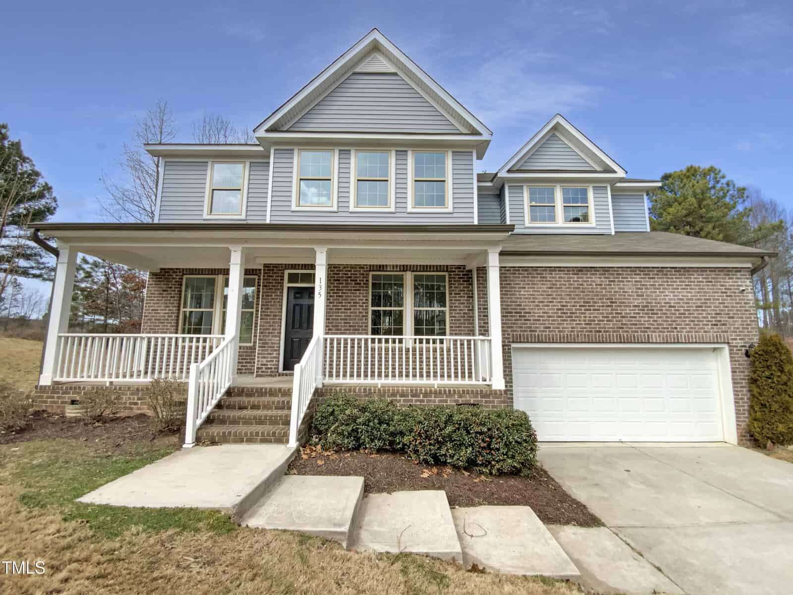 Two-story suburban house with gray siding and brick exterior, white trim, covered front porch with railings, attached two-car garage, concrete driveway, and trimmed bushes in front. Trees and grass yard in the background.