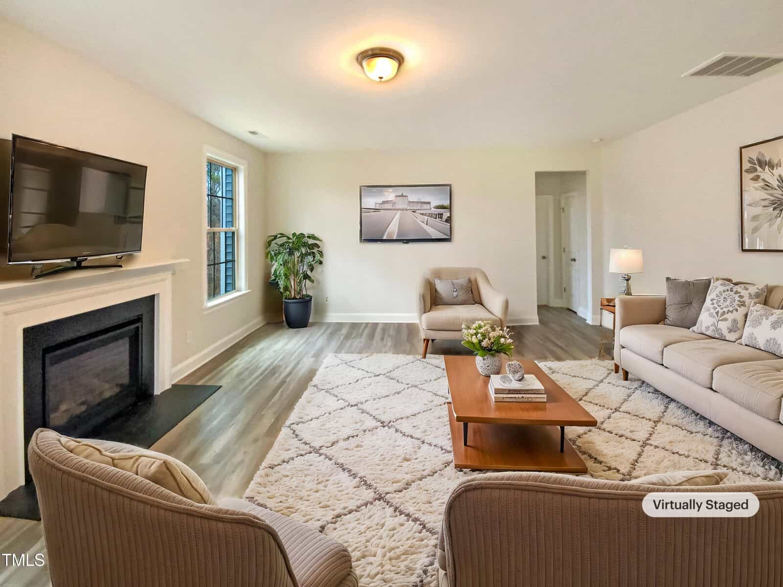 A living room with light walls, wood flooring, a fireplace with a TV above it, a large window, two armchairs, a sofa, a coffee table, a potted plant, wall art, and a rug. The image is virtually staged.