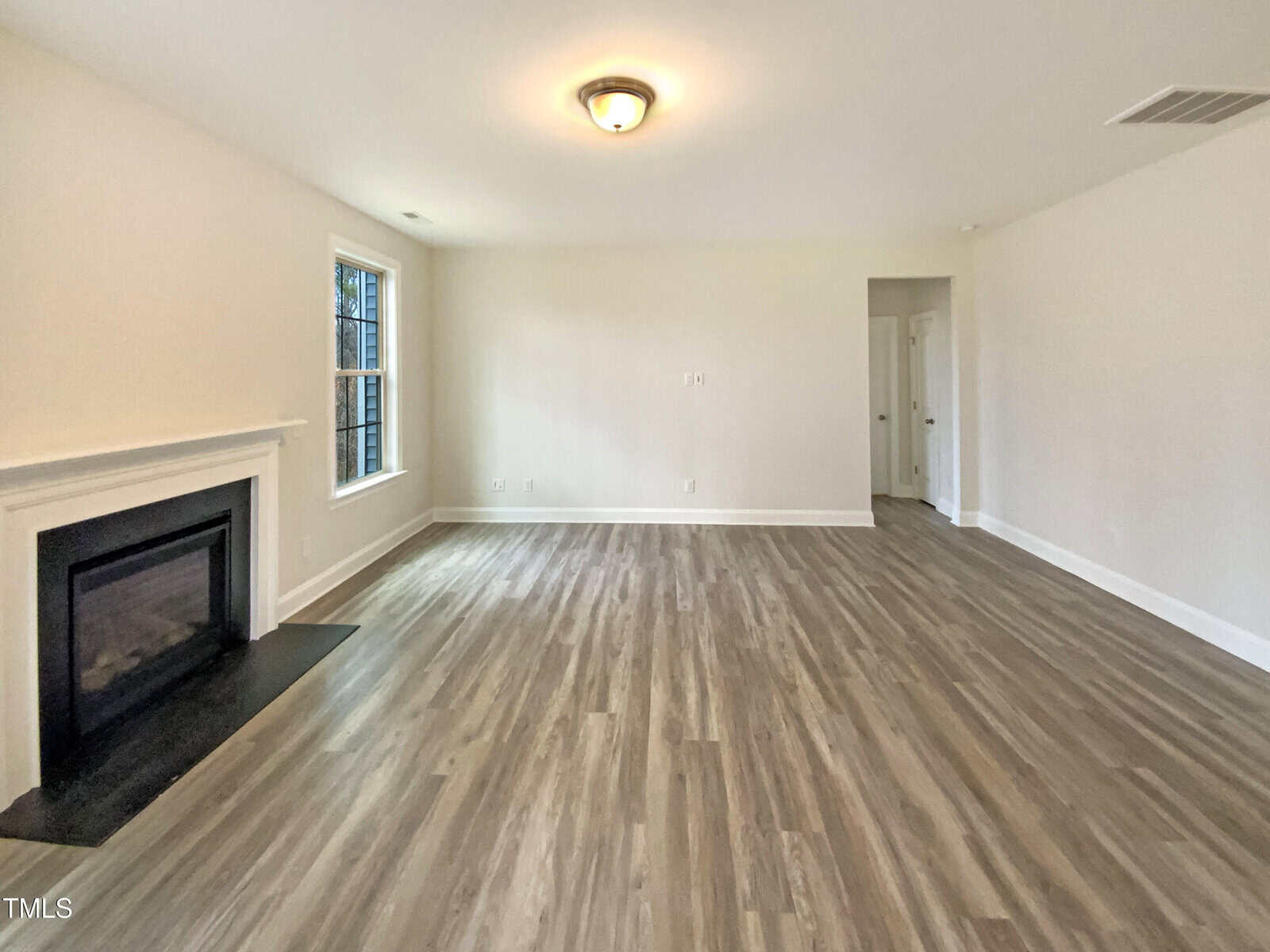 A spacious, empty living room with light-colored walls, wood-style flooring, a black fireplace on the left, a window beside it, and a central ceiling light fixture. An open doorway leads to another area.