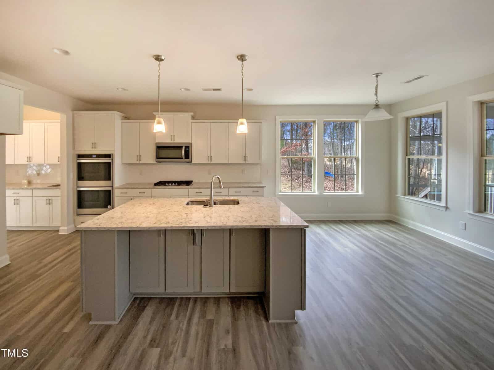 Modern kitchen with white cabinets, double ovens, a center island with sink, pendant lights, and large windows. The space has wood-look flooring and neutral-tone walls, with natural light streaming in through multiple windows.