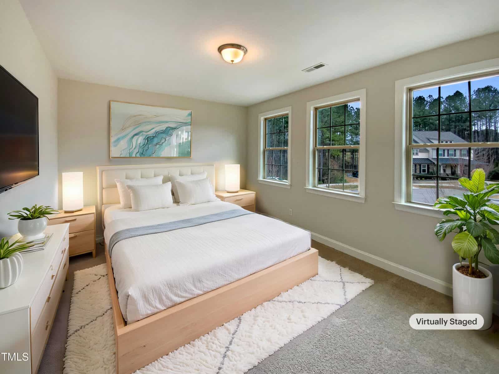 A bedroom with a large bed, two nightstands with lamps, a dresser, a wall-mounted TV, three windows, a potted plant, and artwork above the bed. The room is well-lit and decorated in neutral tones. A “Virtually Staged” label appears.