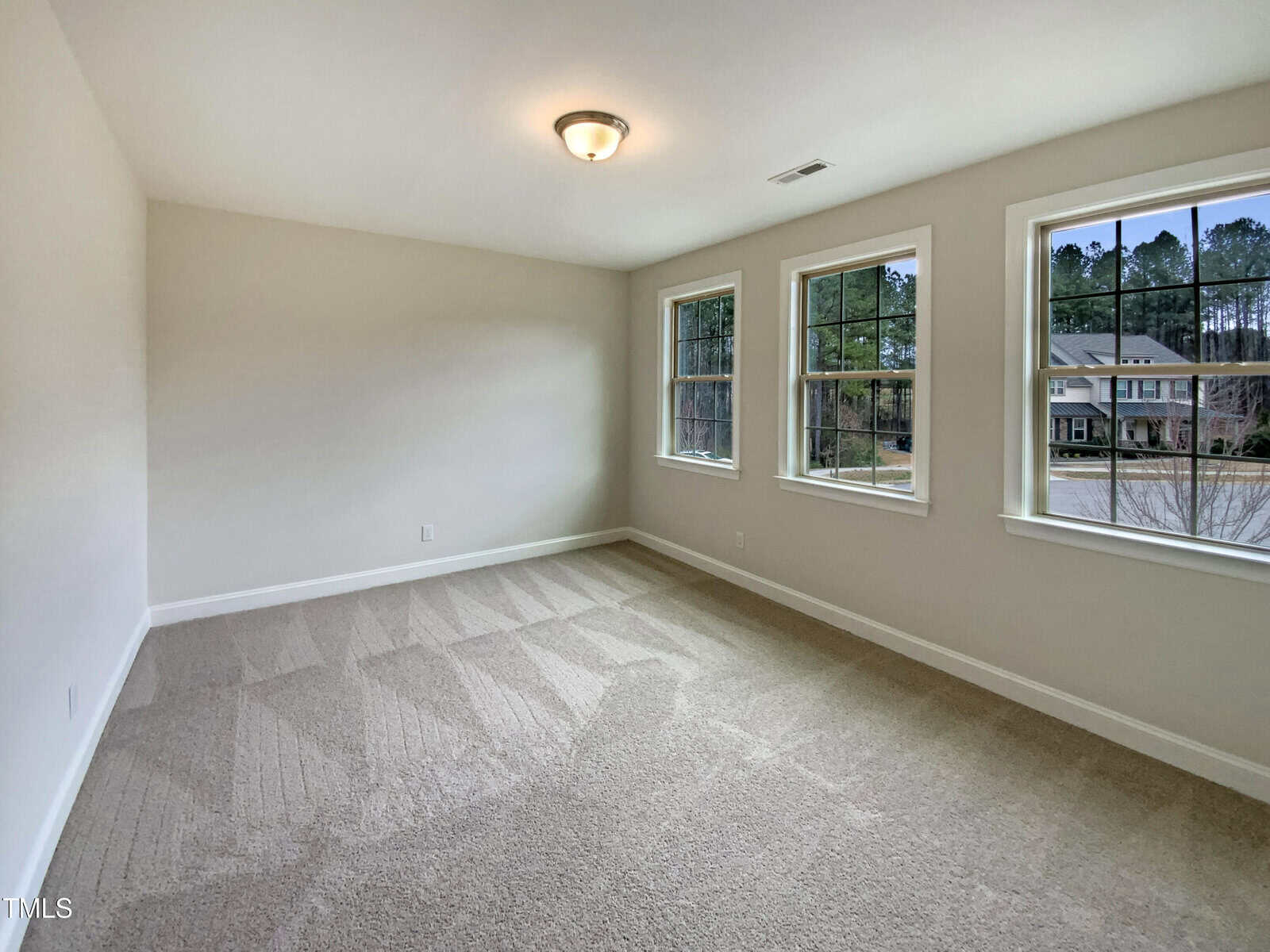 A spacious, empty room with light beige carpet, cream-colored walls, a ceiling light fixture, and three large windows letting in natural light. Trees and houses are visible outside the windows.