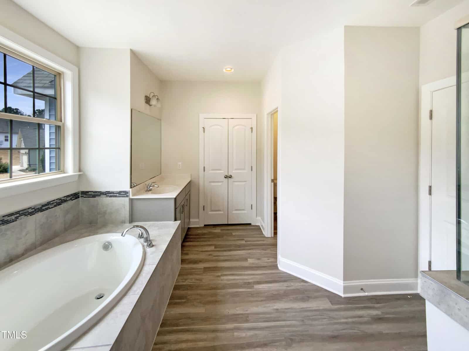 A bathroom with a large bathtub, dual-sink vanity, wood-style flooring, a window, double doors, and a glass shower enclosure in the corner. The walls are light-colored and the room is well-lit.