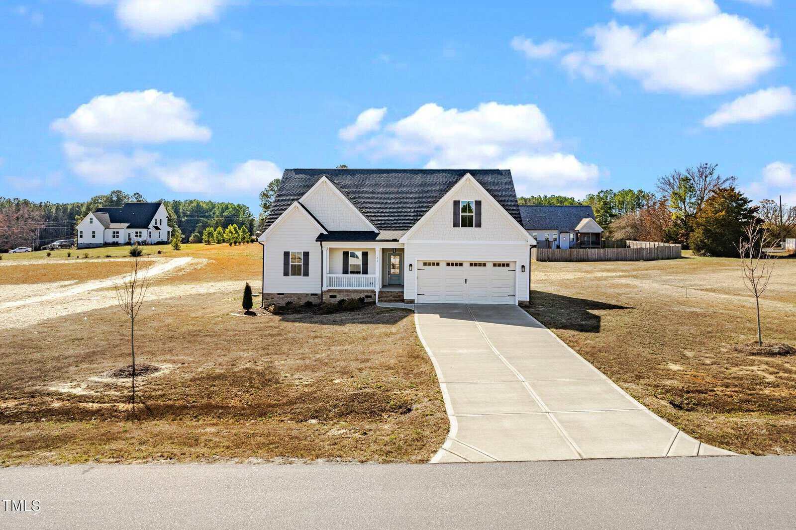 A single-story white house with a two-car garage sits on a large, grassy lot with a long driveway. There are a few young trees in the yard, and another house is visible in the background under a partly cloudy sky.