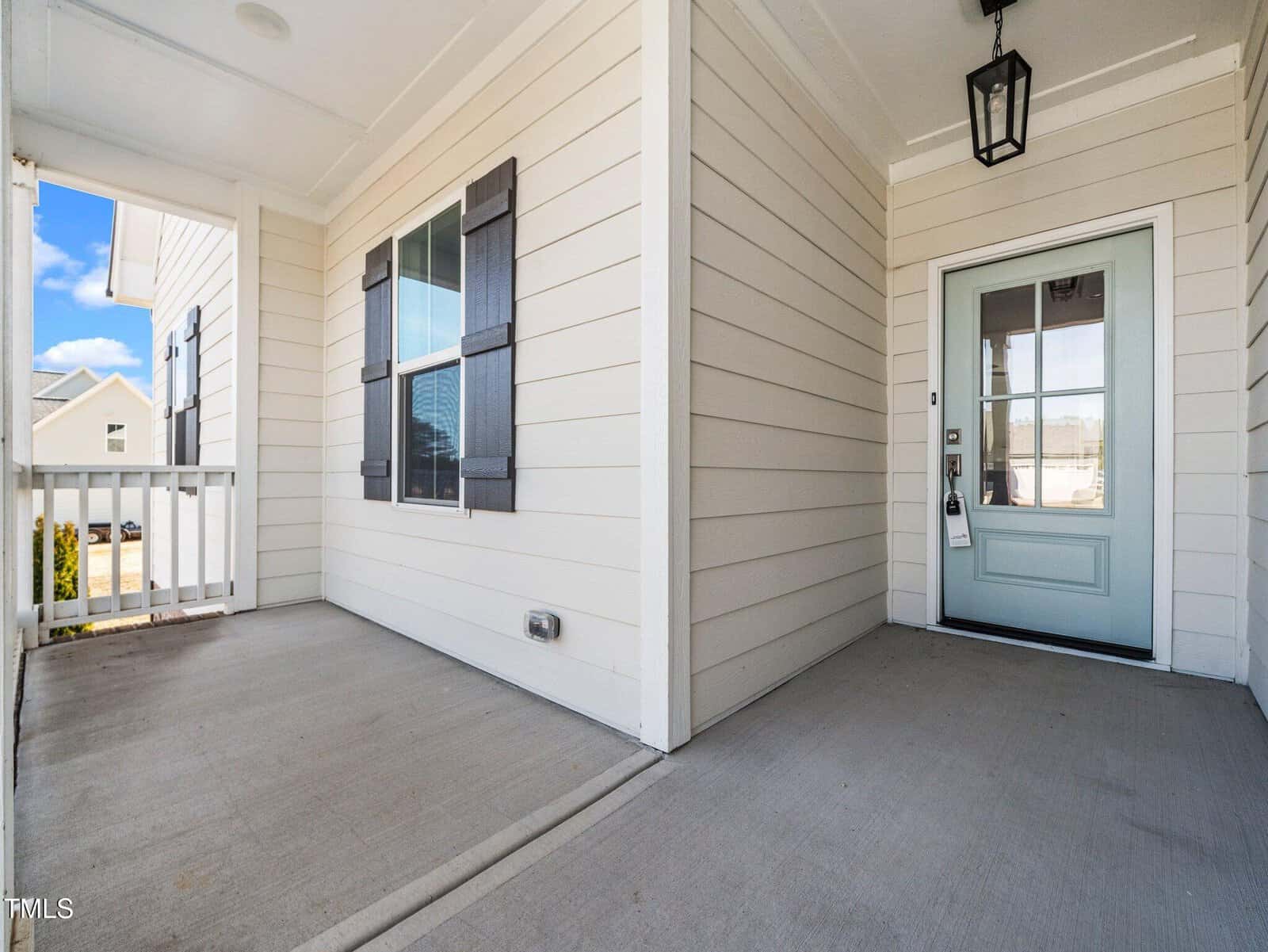 A covered front porch with light-colored siding, two windows with dark shutters, and a light blue front door with glass panels and a black lantern-style light fixture overhead.