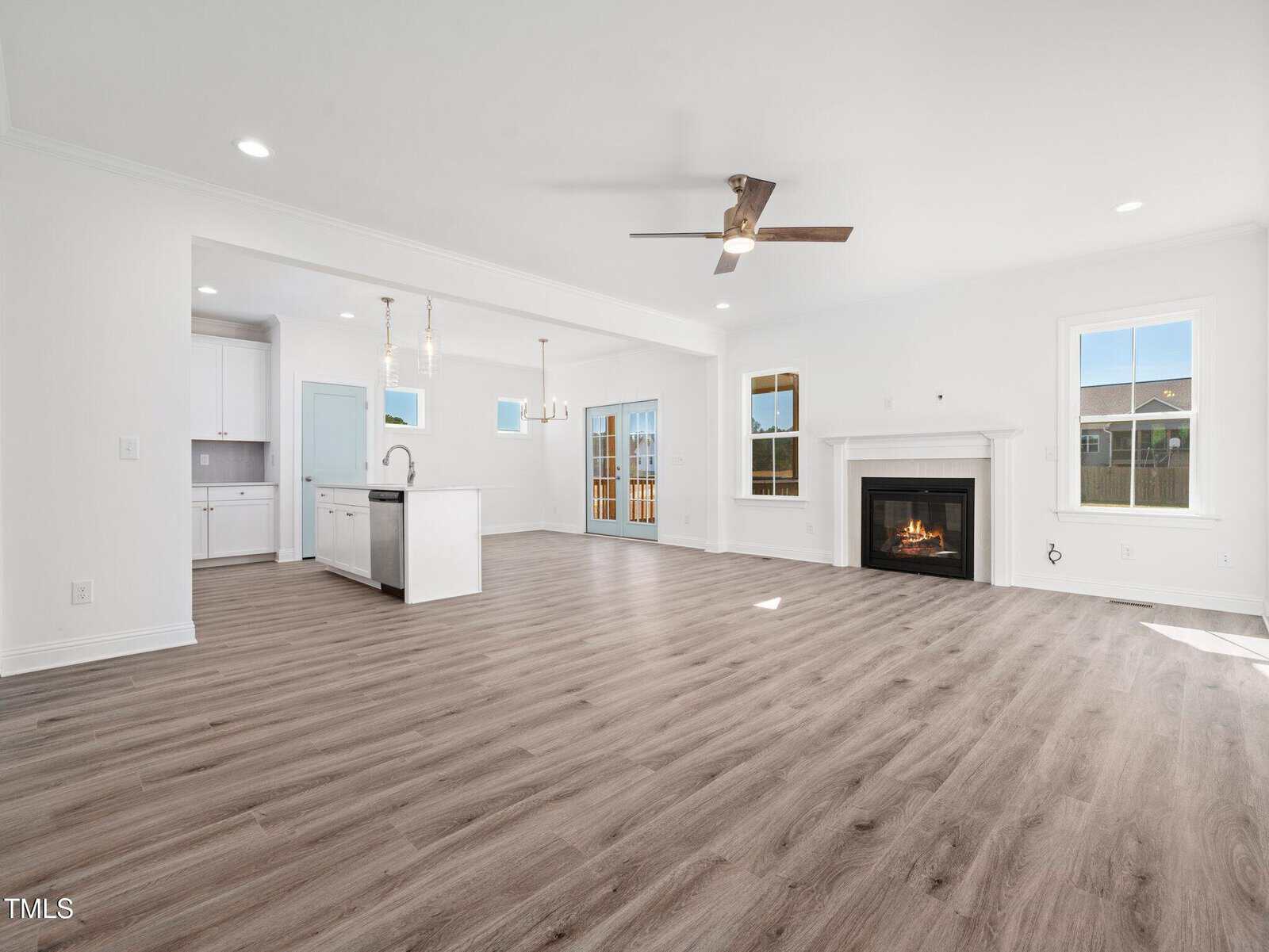Spacious open-concept living area with light wood flooring, white walls, a modern ceiling fan, fireplace, large windows, and an adjacent kitchen featuring an island, white cabinetry, and pendant lights.