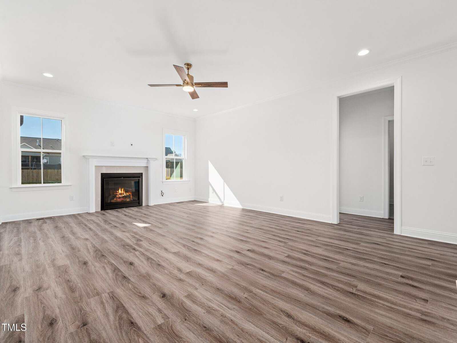 A spacious living room with light wood flooring, white walls, a ceiling fan, recessed lighting, a fireplace with a white mantel, and two windows allowing natural light into the space.