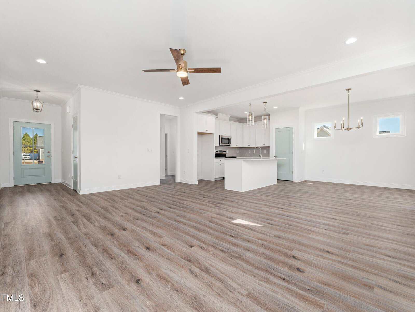 Spacious open-concept living area with light wood flooring, white walls, a ceiling fan, modern kitchen with island and stainless steel appliances, and natural light from several windows.