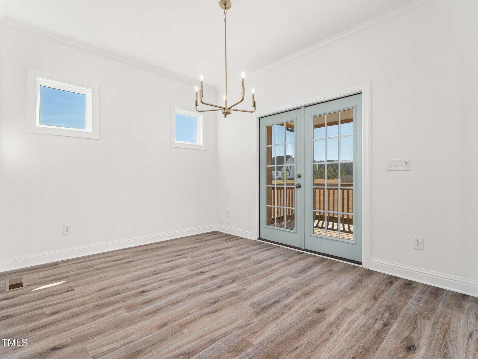 Bright, empty room with light wood flooring, white walls, two small windows, a modern chandelier, and double glass doors leading to a wooden deck outside. Natural light enters through windows and doors.