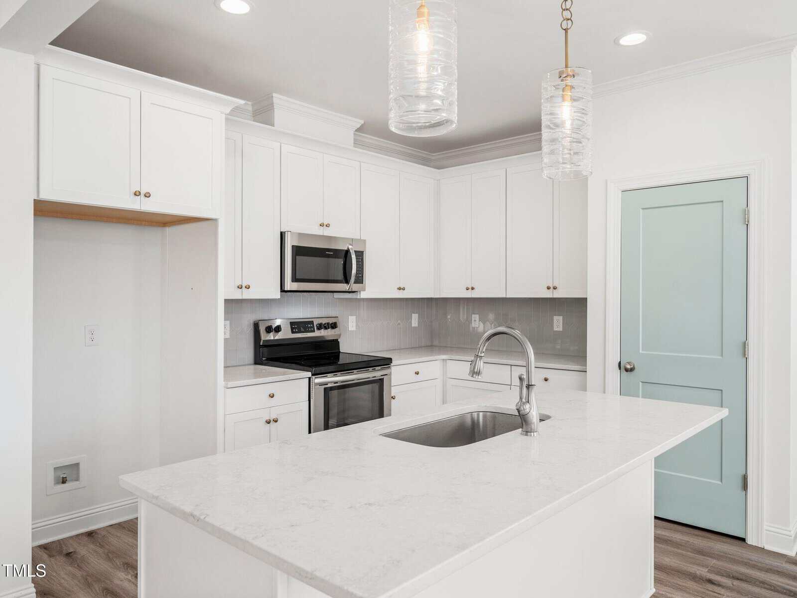 Modern kitchen with white cabinets, stainless steel appliances, a light blue door, white countertops, and pendant lights above an island with a sink. The room has light wood flooring and light gray tile backsplash.