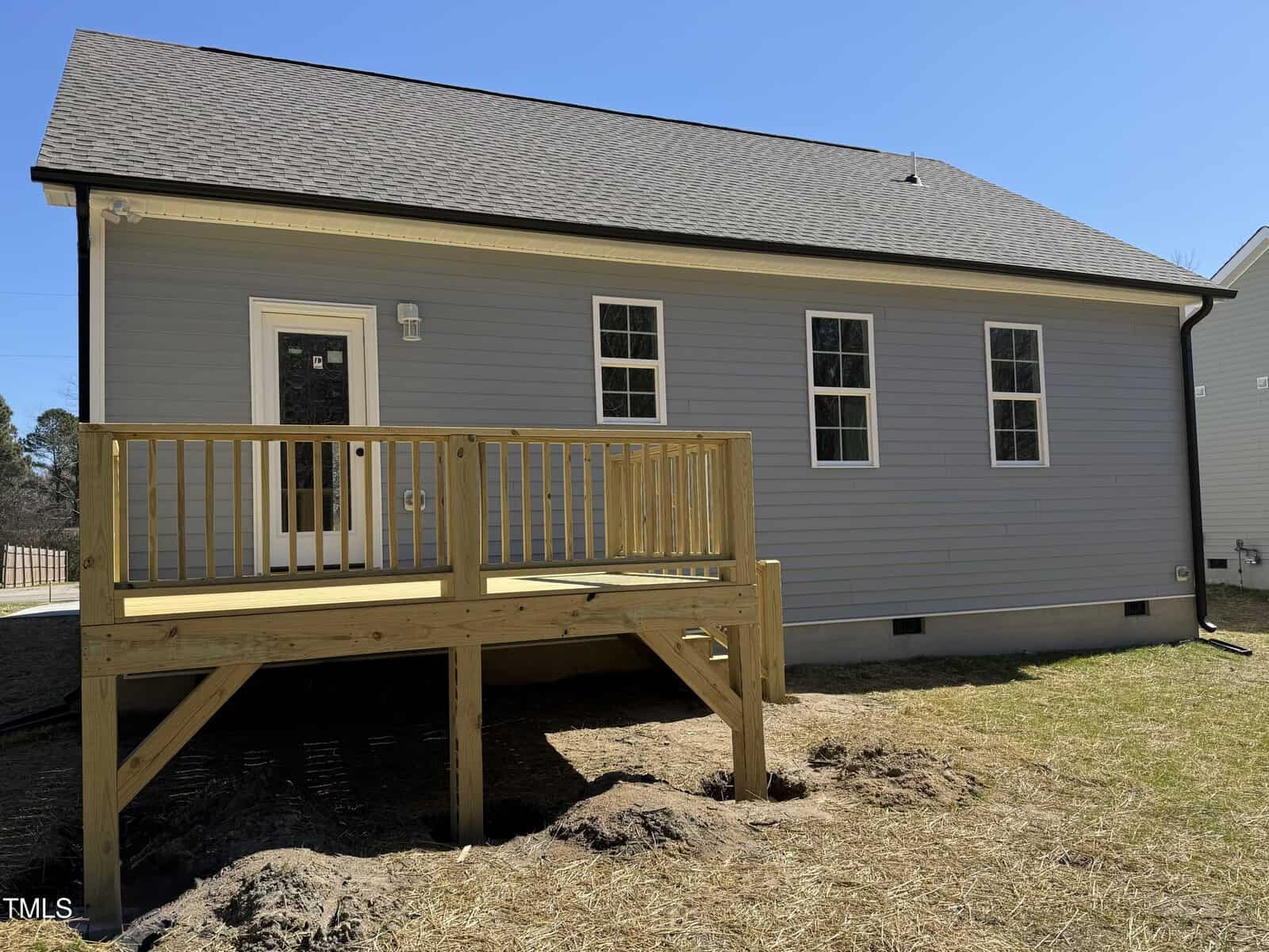 Single-story gray house with a sloped roof and three windows. There is a small wooden deck with railings and three steps leading down to a grassy backyard. A glass door provides access to the deck from the house.