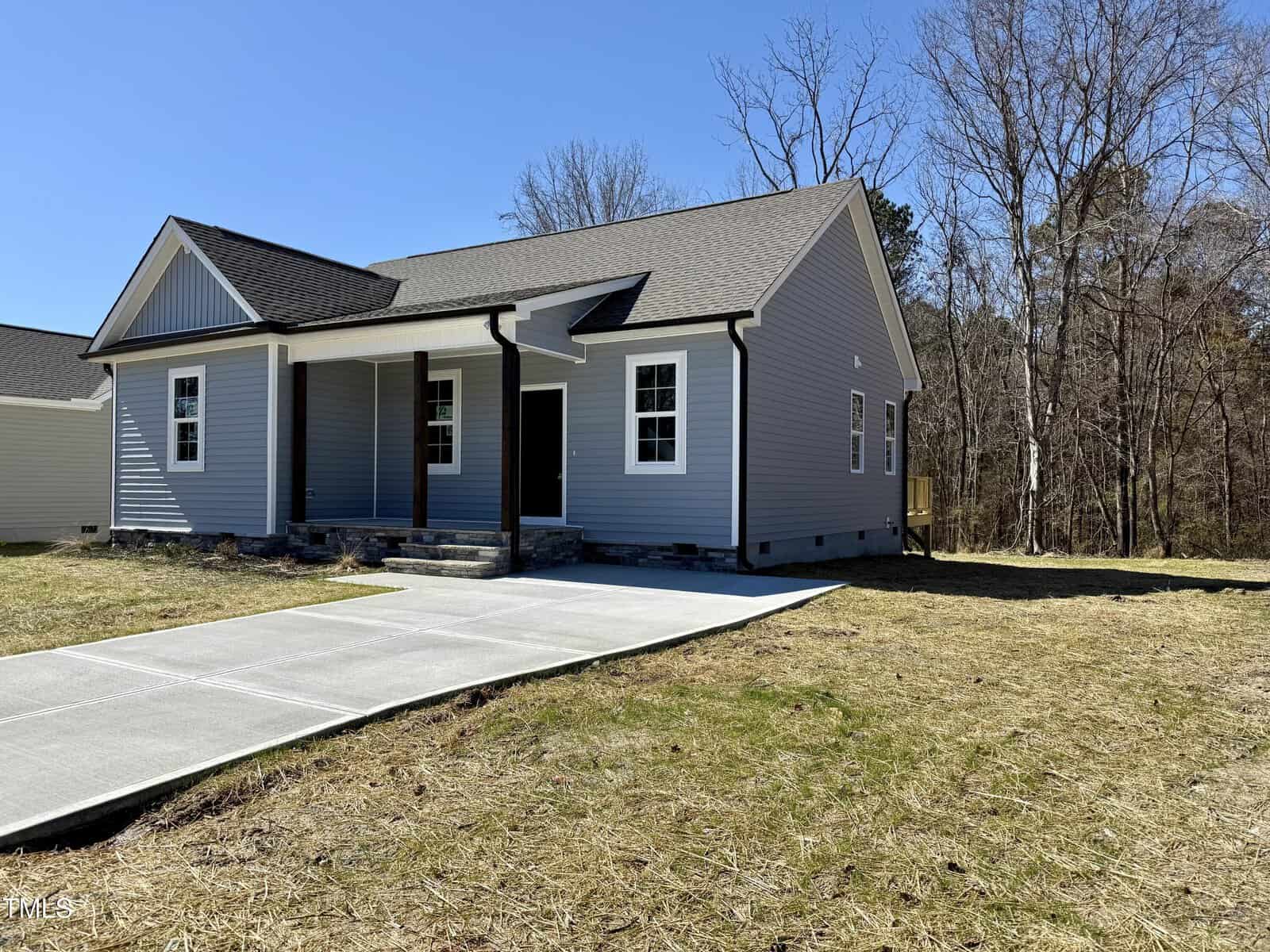 A gray single-story house with white trim and a gabled roof, featuring a small front porch supported by dark wooden columns. A concrete driveway leads to the entrance. The house is surrounded by a grassy area and trees in the background.