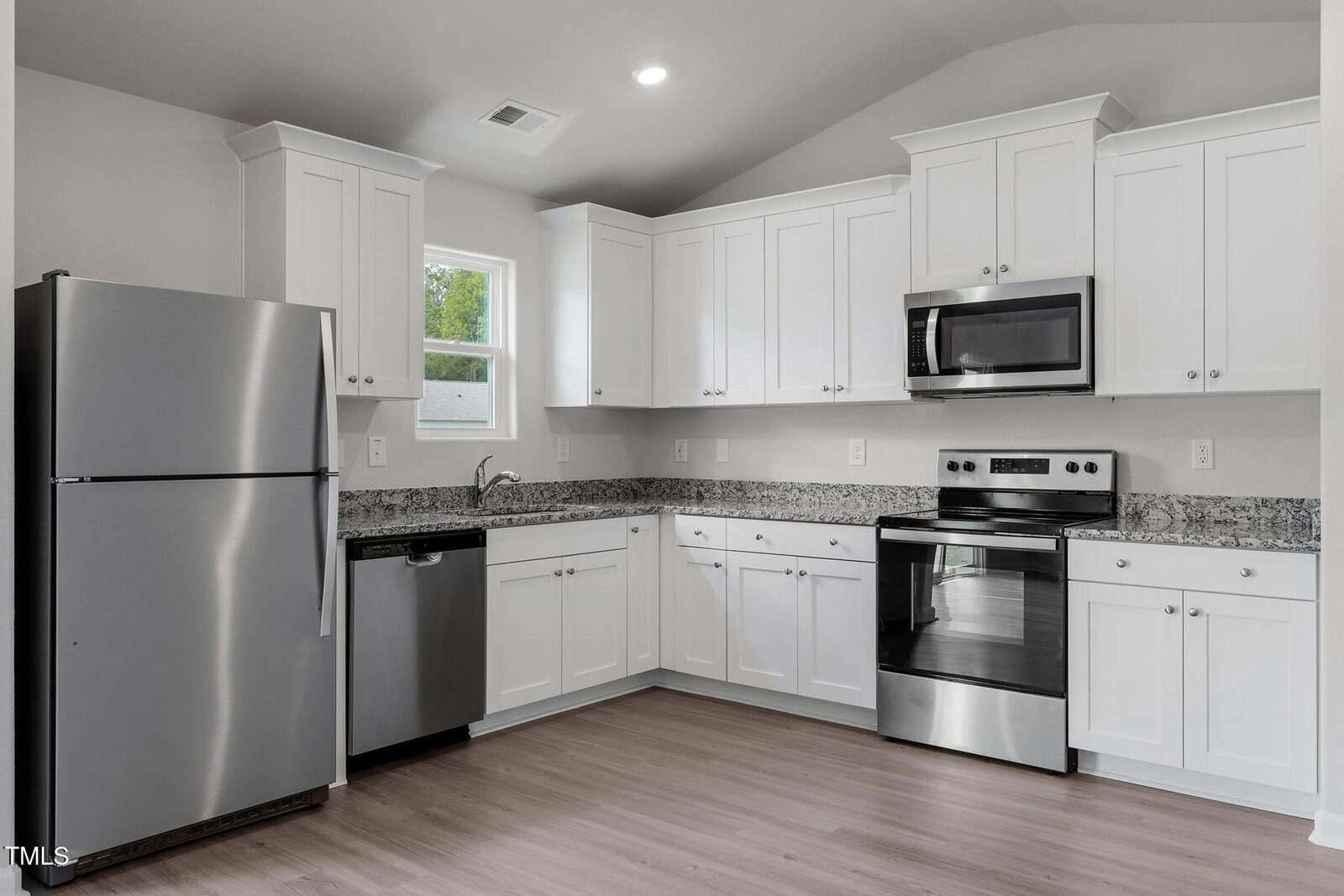 Modern kitchen with white cabinets, stainless steel appliances including a refrigerator, dishwasher, and oven with a stove. Granite countertops and light wooden flooring. A small window is above the sink, providing natural light.