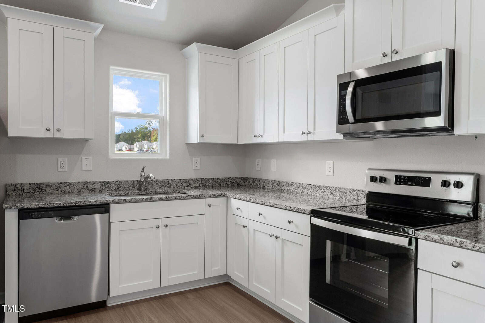 Modern kitchen with white cabinets and granite countertops. Stainless steel appliances include a dishwasher, microwave, and oven. A small window above the sink lets in natural light. Wood flooring contrasts with the white and gray decor.