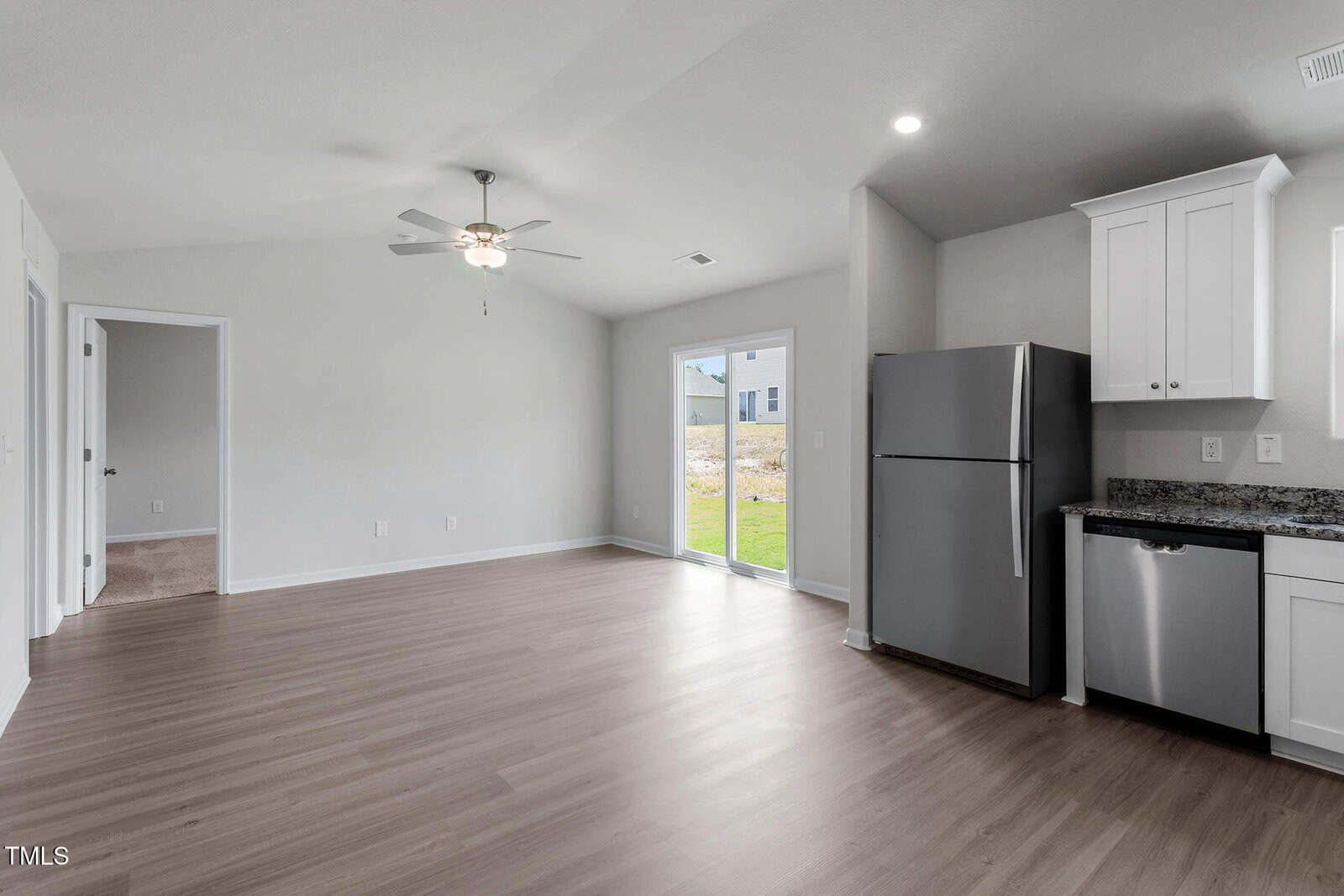 A modern, unfurnished kitchen and living area with light wood flooring. It includes white cabinets, a stainless steel refrigerator, and dishwasher. A ceiling fan is visible above, and there is a large windowed door leading outside.