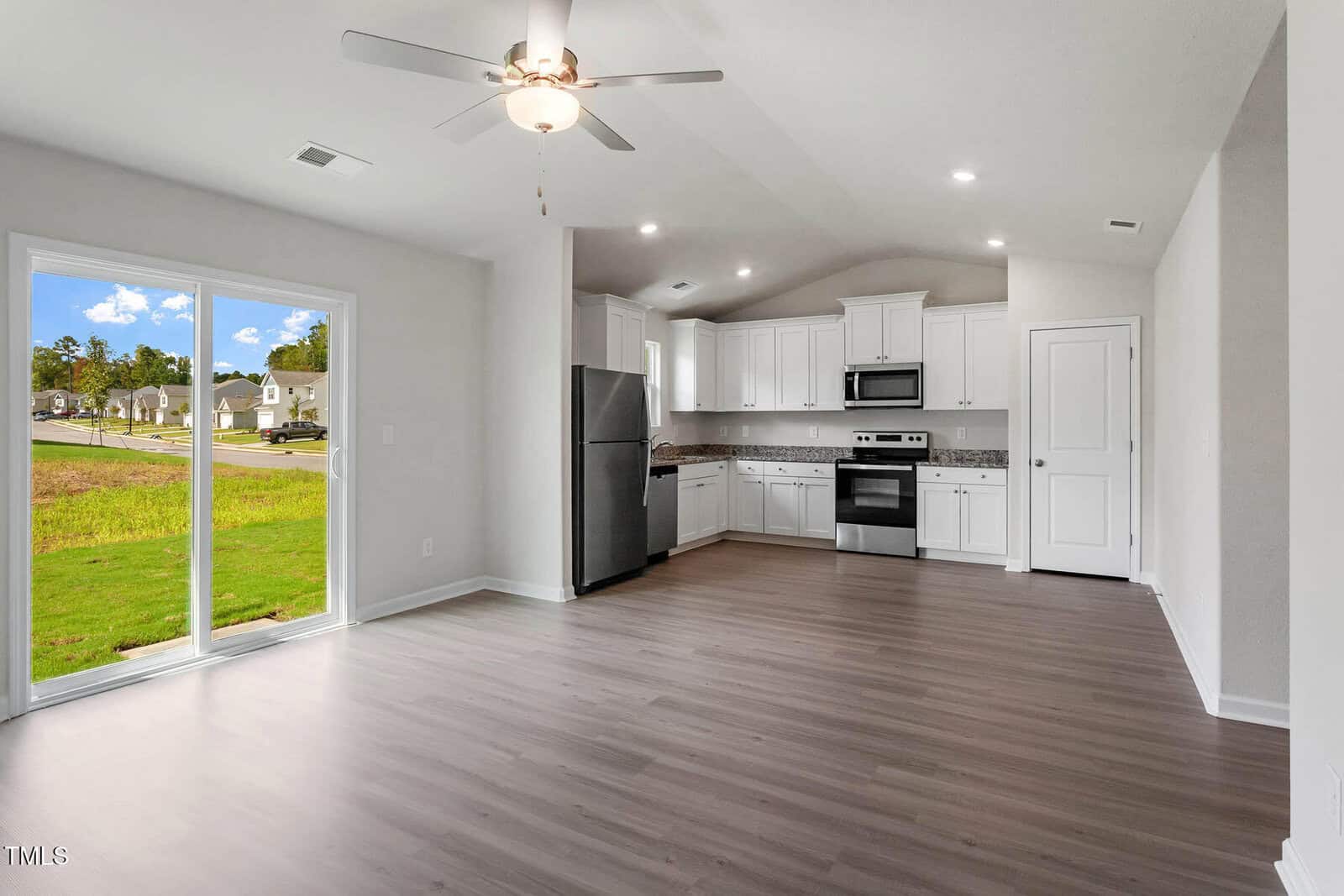 A modern kitchen and living area with light wood flooring. The kitchen features white cabinets, stainless steel appliances, and a granite countertop. A ceiling fan is mounted above. Large sliding glass doors open to a grassy outdoor area with houses visible.