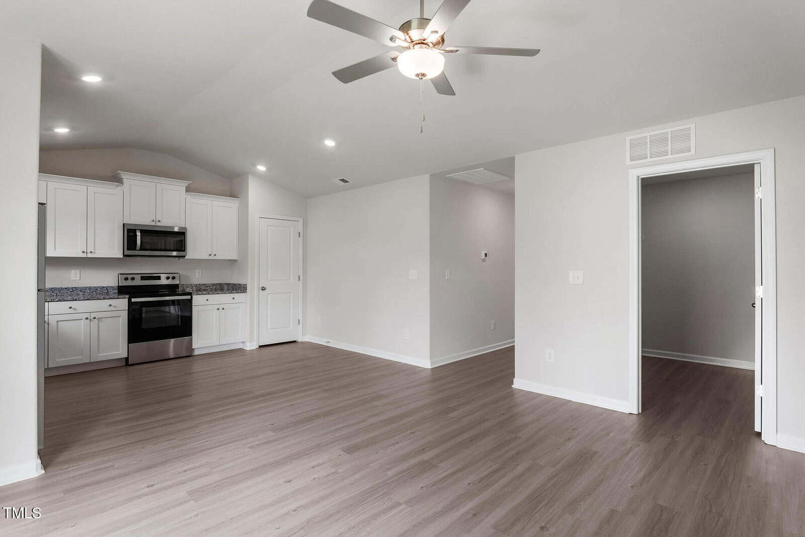 Spacious empty room with light wood flooring and white walls. A ceiling fan is centered in the room. The kitchen area features white cabinets, stainless steel appliances, and granite countertops. An open doorway leads to another room.