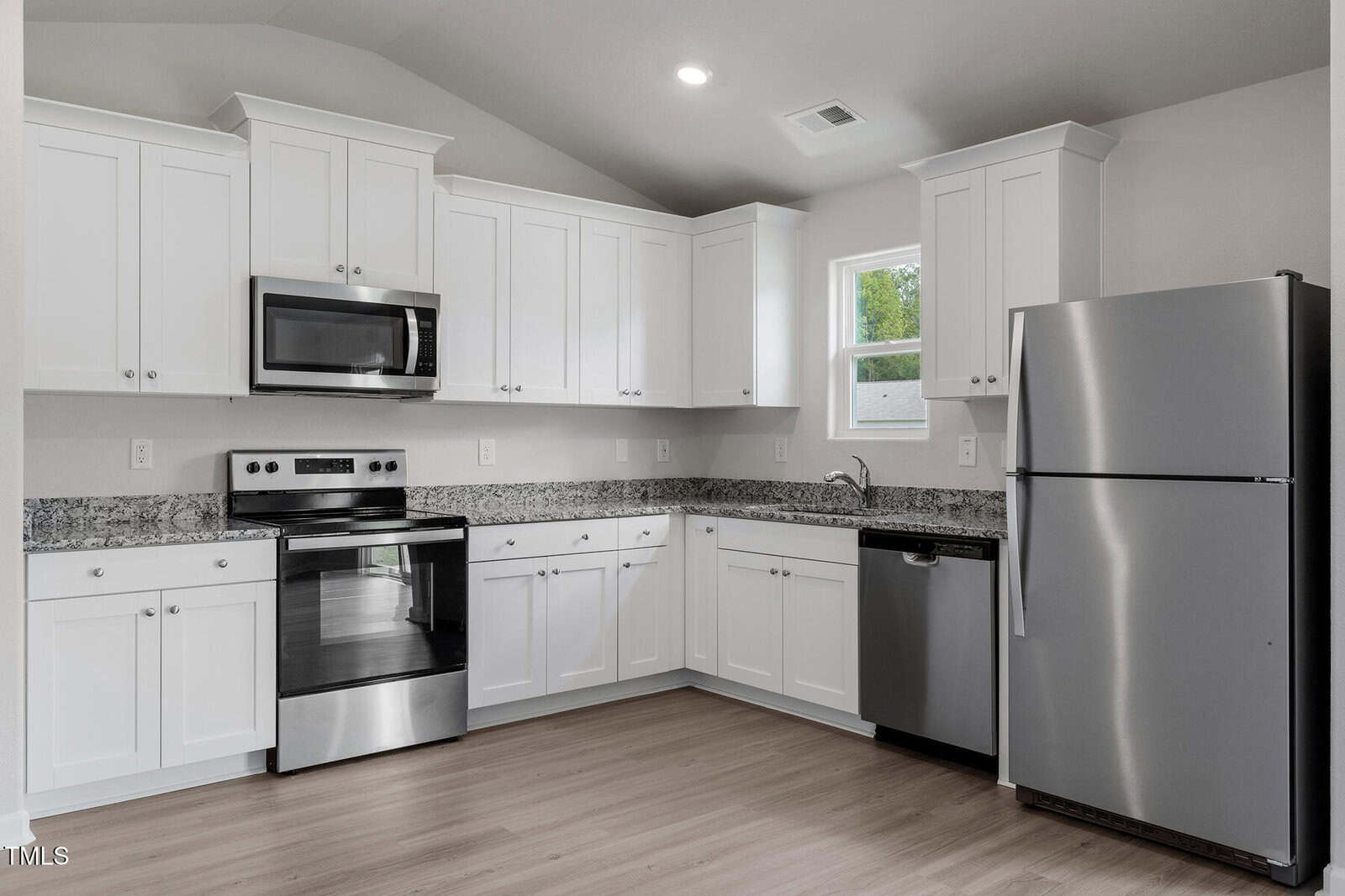 Modern kitchen with white cabinetry, stainless steel appliances including a refrigerator, oven, microwave, and dishwasher. Granite countertops and light wood flooring are present, with a small window above the sink area.