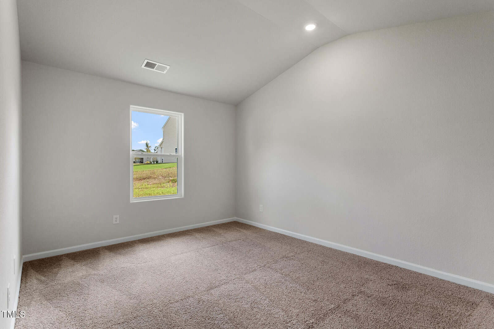 An empty room with beige carpet and light gray walls, featuring a single window with a view of grass and part of a house. The ceiling is vaulted with a recessed light fixture.